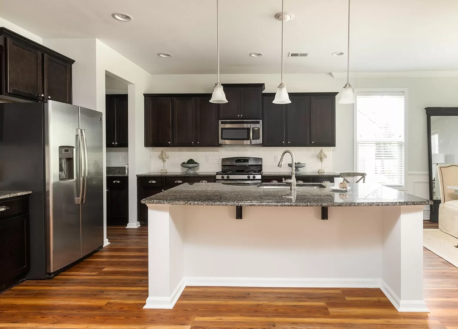 Modern kitchen with dark cabinets, stainless steel refrigerator and microwave, granite countertops, white walls, hardwood floors, and pendant lighting above the island.