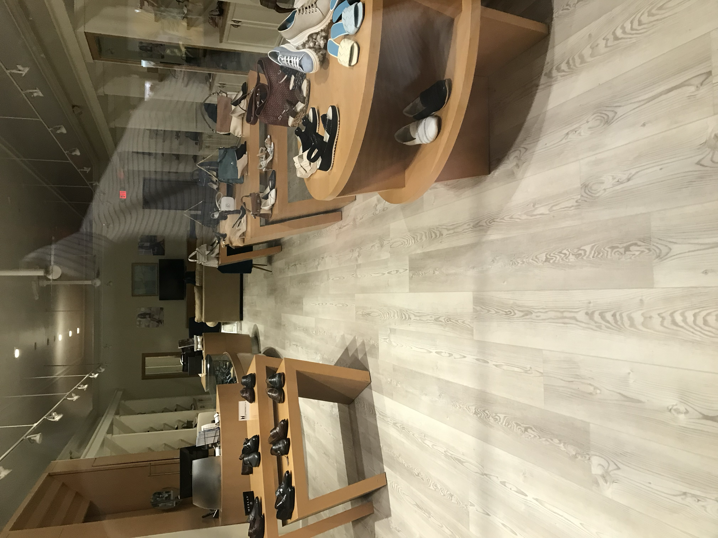 A retail store displaying shoes and handbags on wooden tables and shelves, with sofas and television in the background.