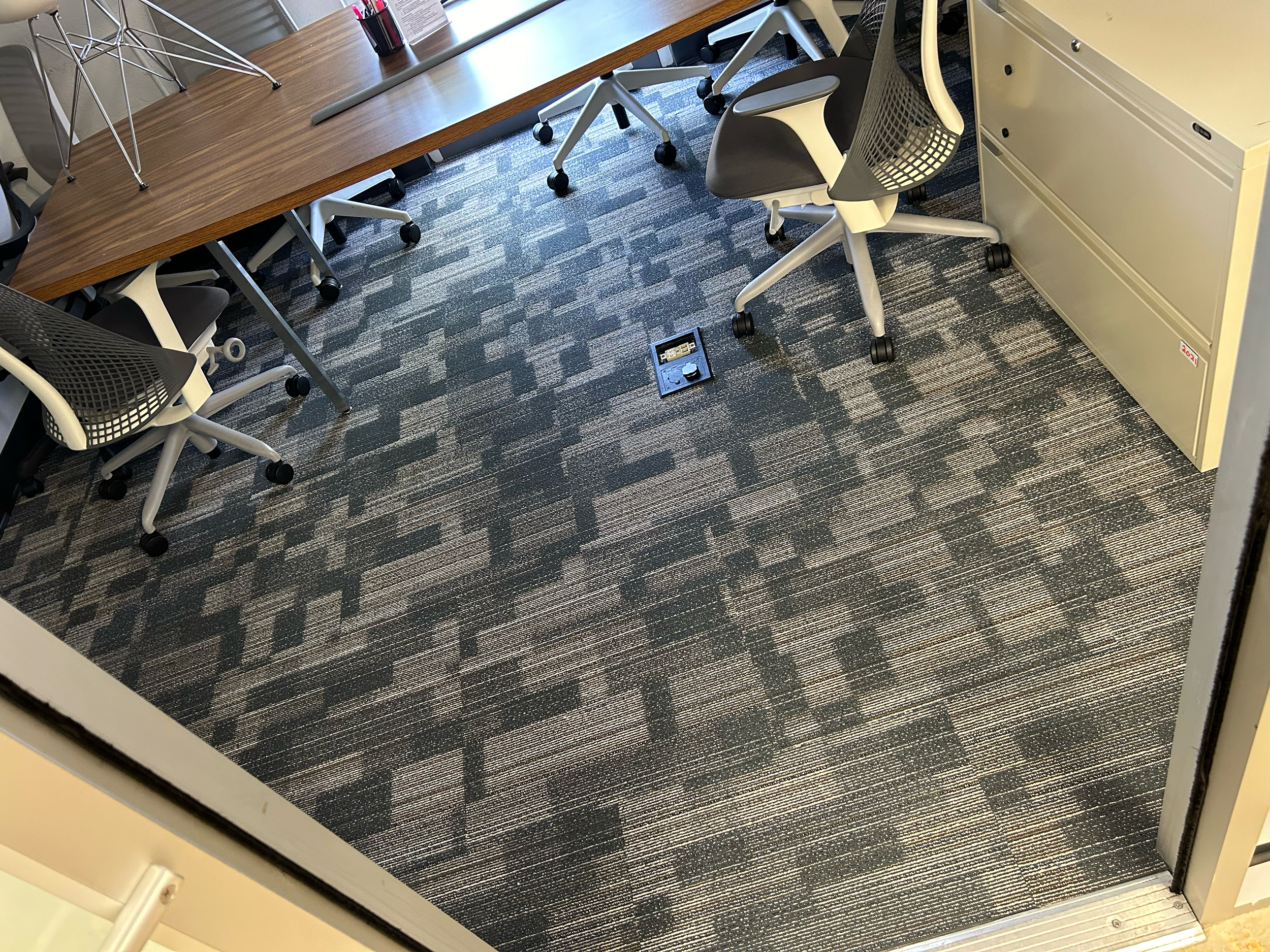 Office room with a large wooden conference table, several gray and white office chairs on wheels, a window with blinds, a white filing cabinet, a metal electrical socket on the carpeted floor, and a metal sculpture on the table.