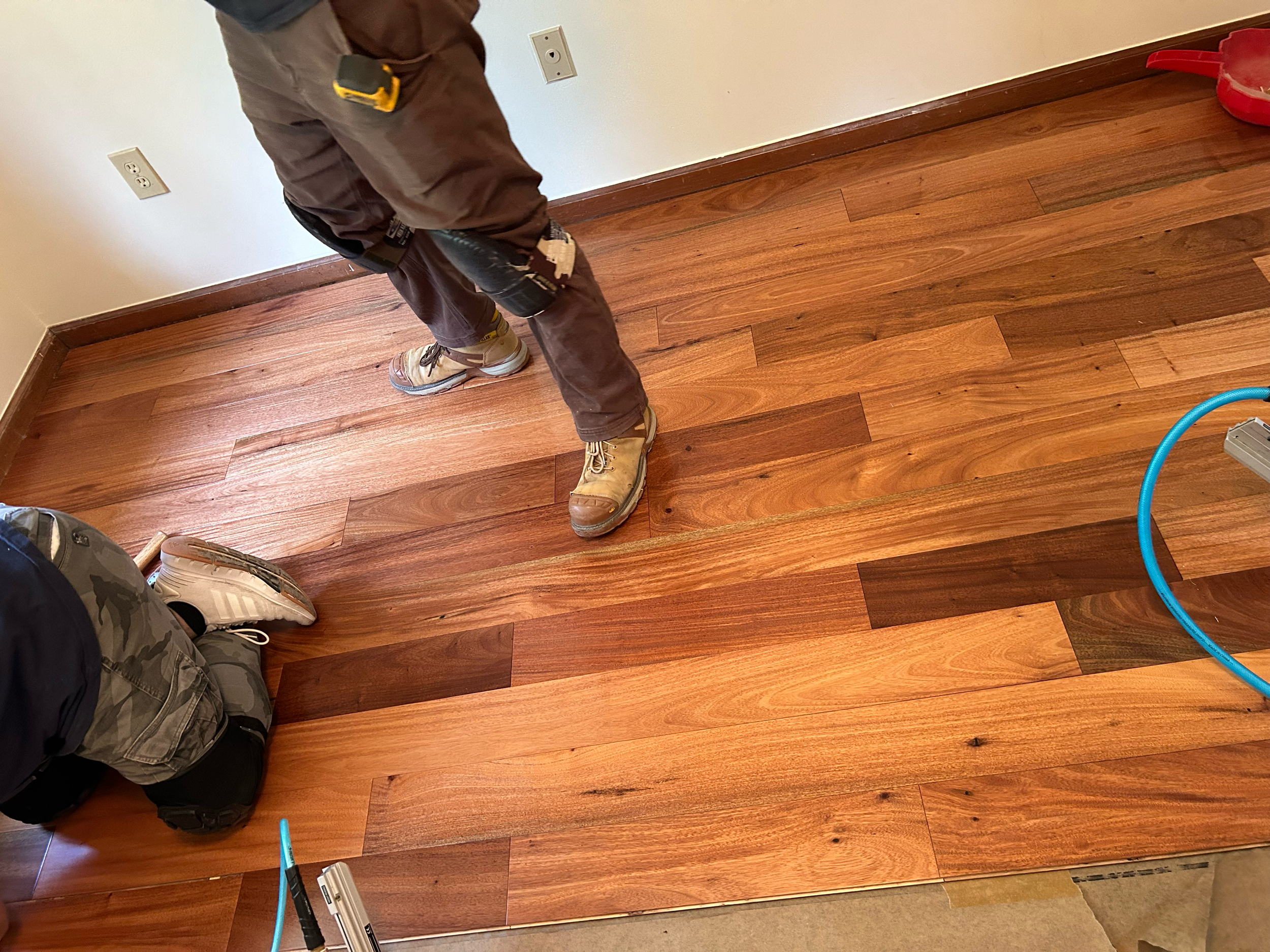 Person installing hardwood flooring, holding a power tool, with a section of completed wooden floor visible.