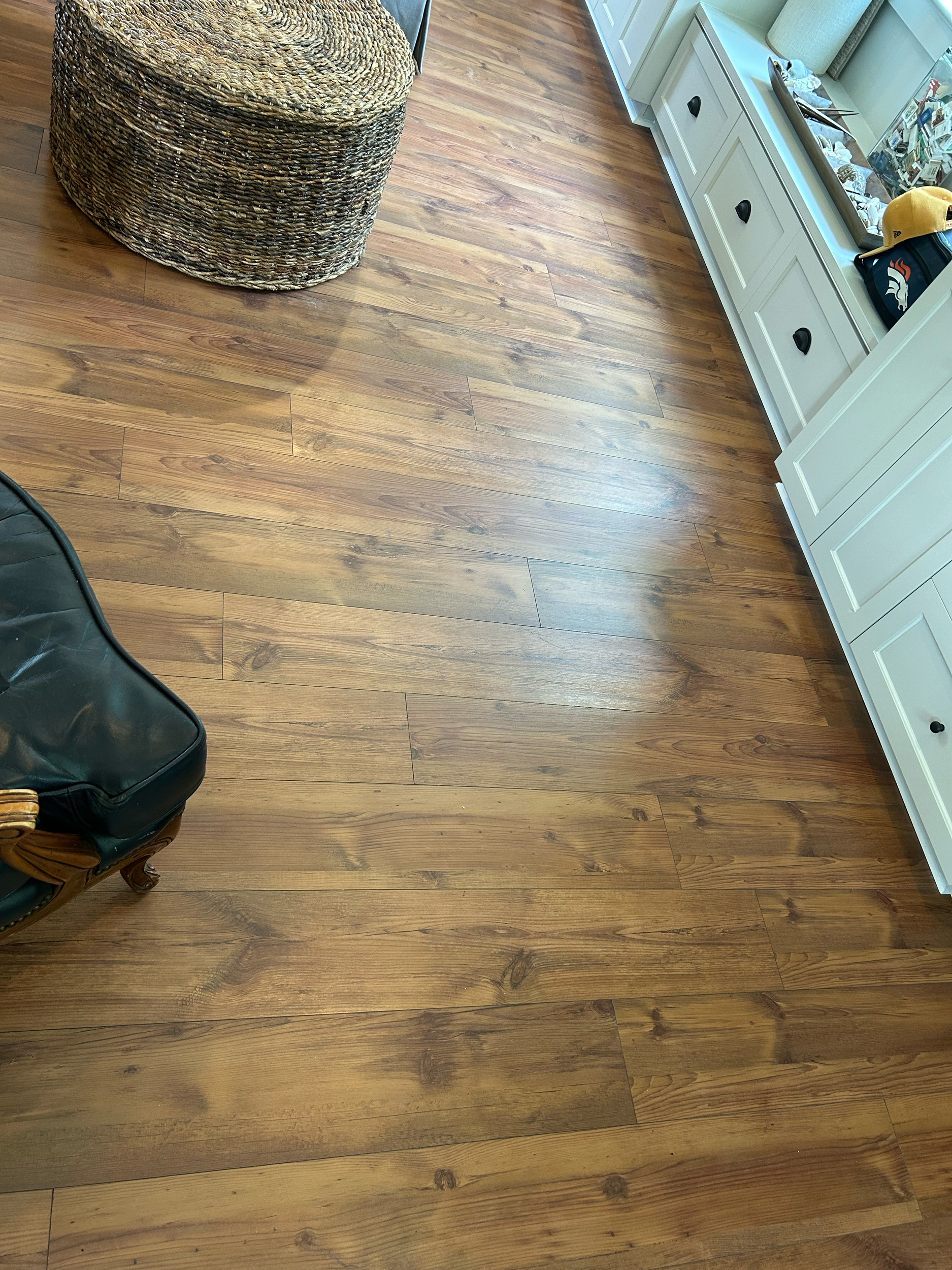 A room with wooden flooring, a wicker ottoman, a white cabinet with drawers, and some items on top including a bag and a hat.