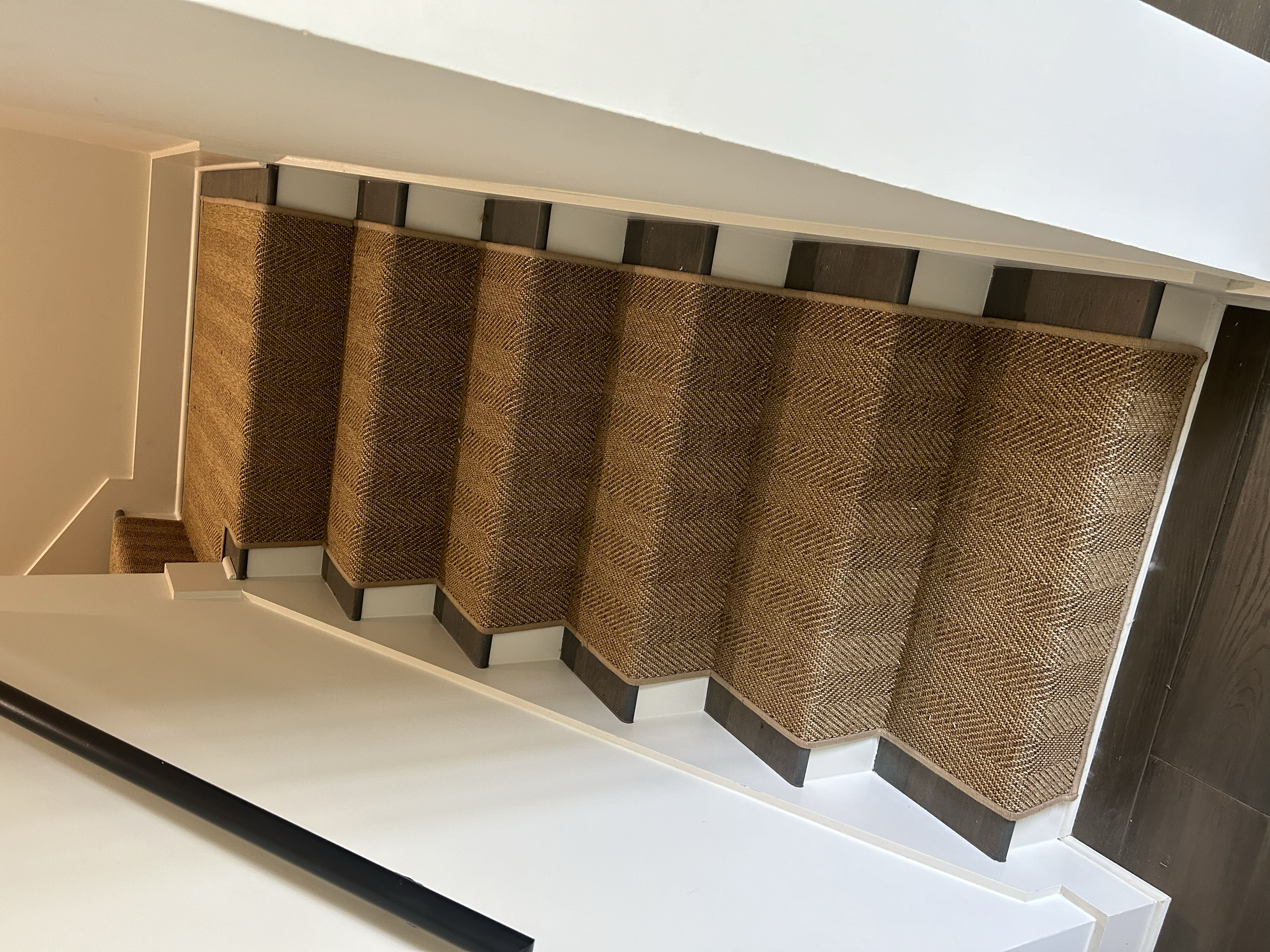 A staircase with four steps covered by a brown patterned carpet, with white walls on either side and a dark wooden floor at the bottom.