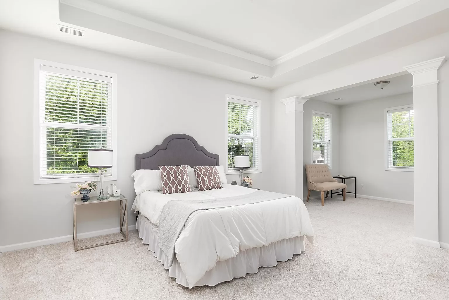 Bright, modern bedroom with white walls, large windows, a bed with a tufted gray headboard, white bedding, decorative pillows, side tables with lamps, a beige armchair, and a small black table.