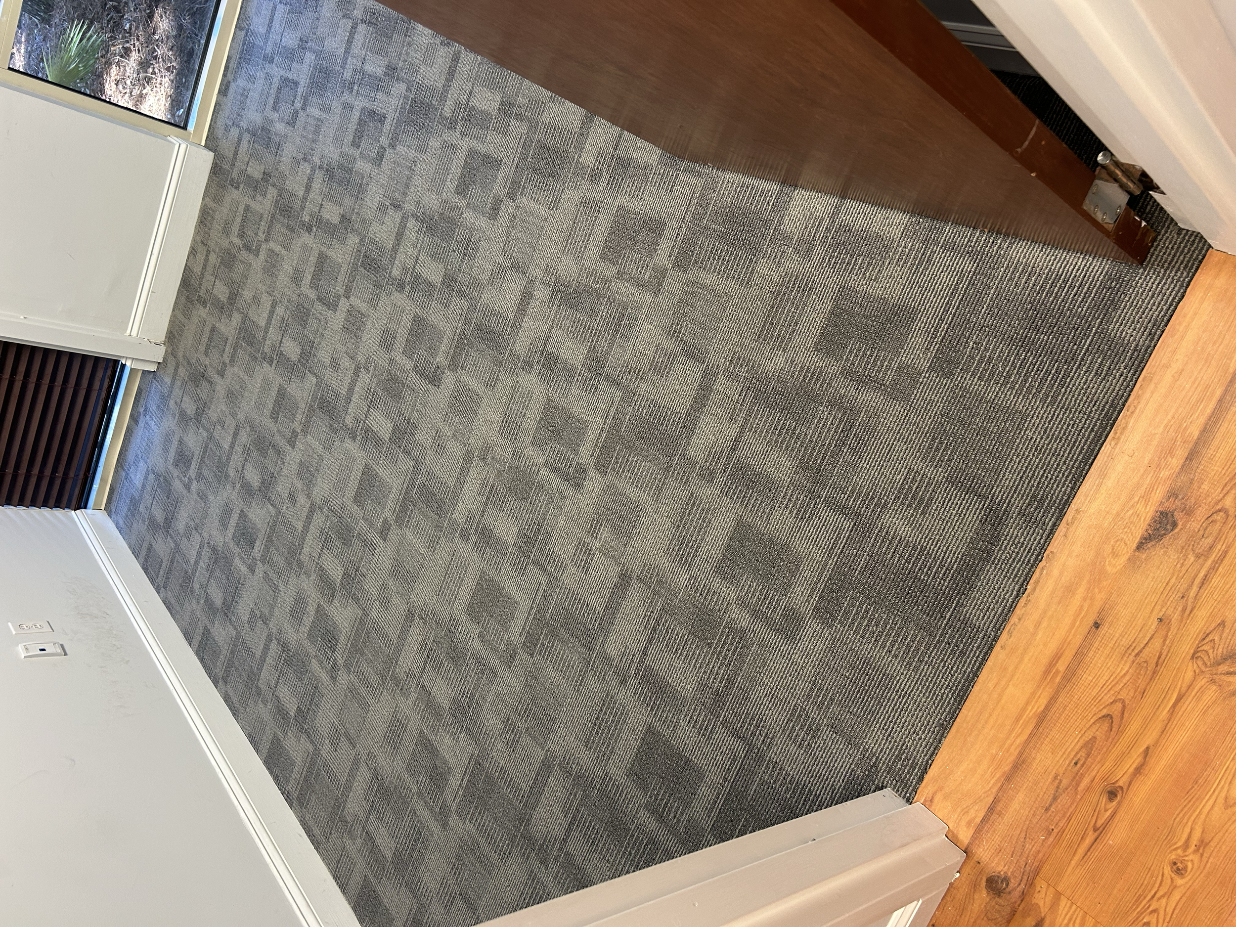Photo of a room entryway with a grey patterned carpet, a wooden floor, white walls, electrical outlets, a window with brown blinds, and a door with a silver handle.