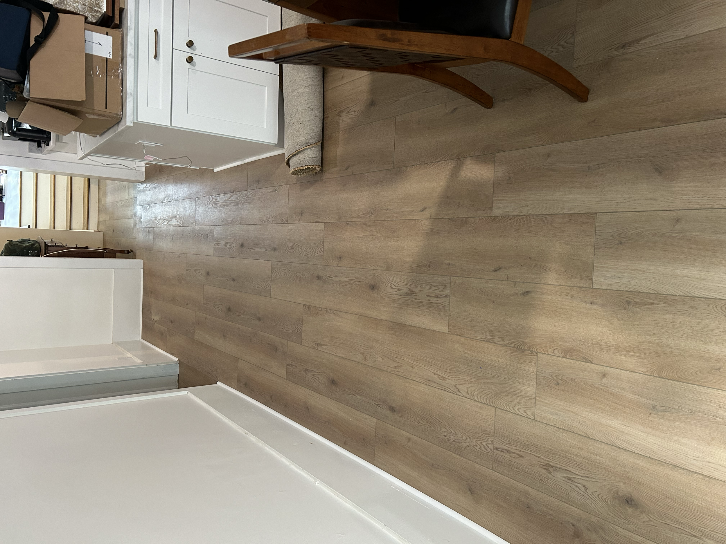 Wooden flooring in a home interior with a doorway leading to stairs, a white cabinet with a cardboard box on top, and a rolled-up carpet or rug on the floor.