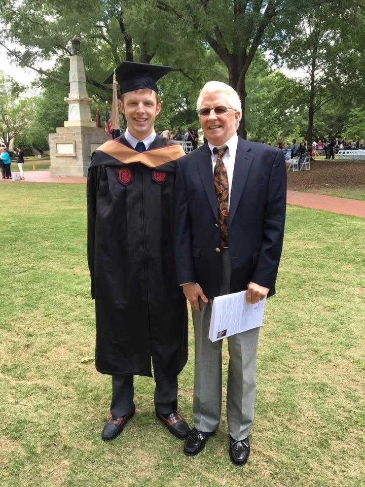 A young man in a graduation cap and gown standing next to an older man in a suit during a graduation ceremony outdoors.