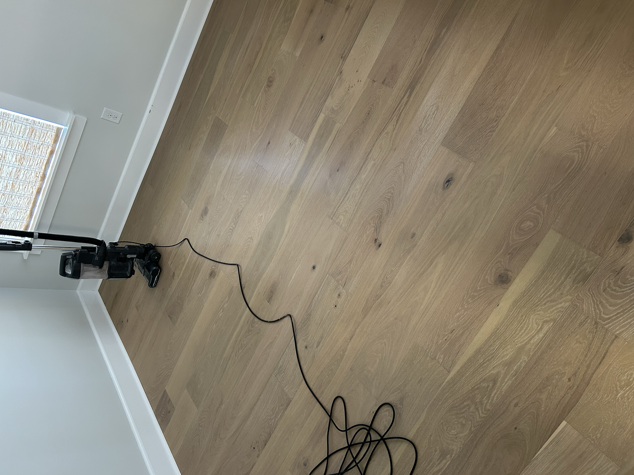 A black upright vacuum cleaner with a black power cord on a light hardwood floor near a corner of a room with a white baseboard and a window with blinds.