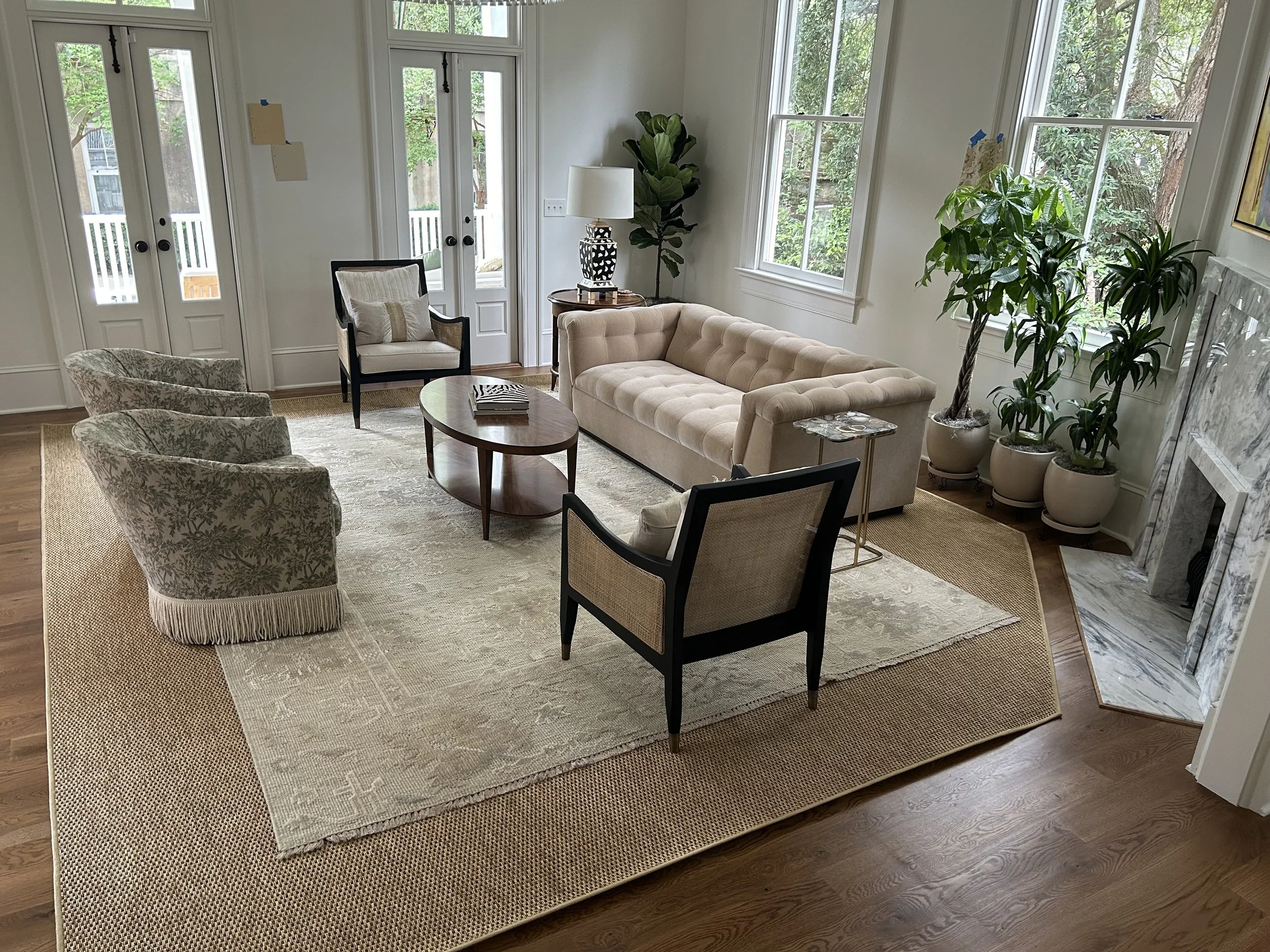 A living room with a beige tufted sofa, two patterned armchairs, a wooden coffee table, large windows with green trees outside, a potted plant near the door, and a rug on hardwood floor.