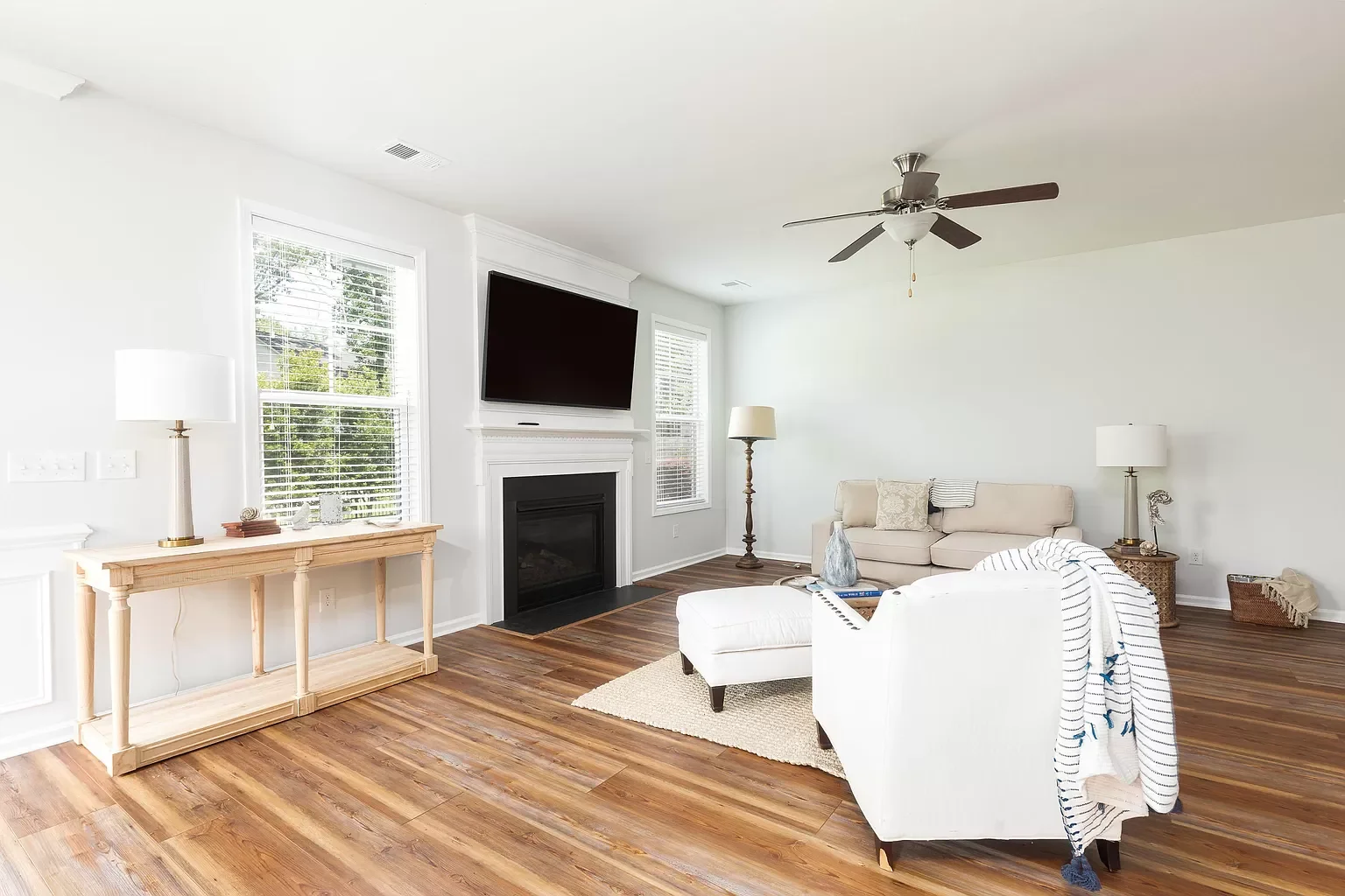 Living room with hardwood floor, white walls, large windows, a wall-mounted TV above a fireplace, a beige sofa, white armchair with a striped blanket, wooden side tables, and multiple lamps.