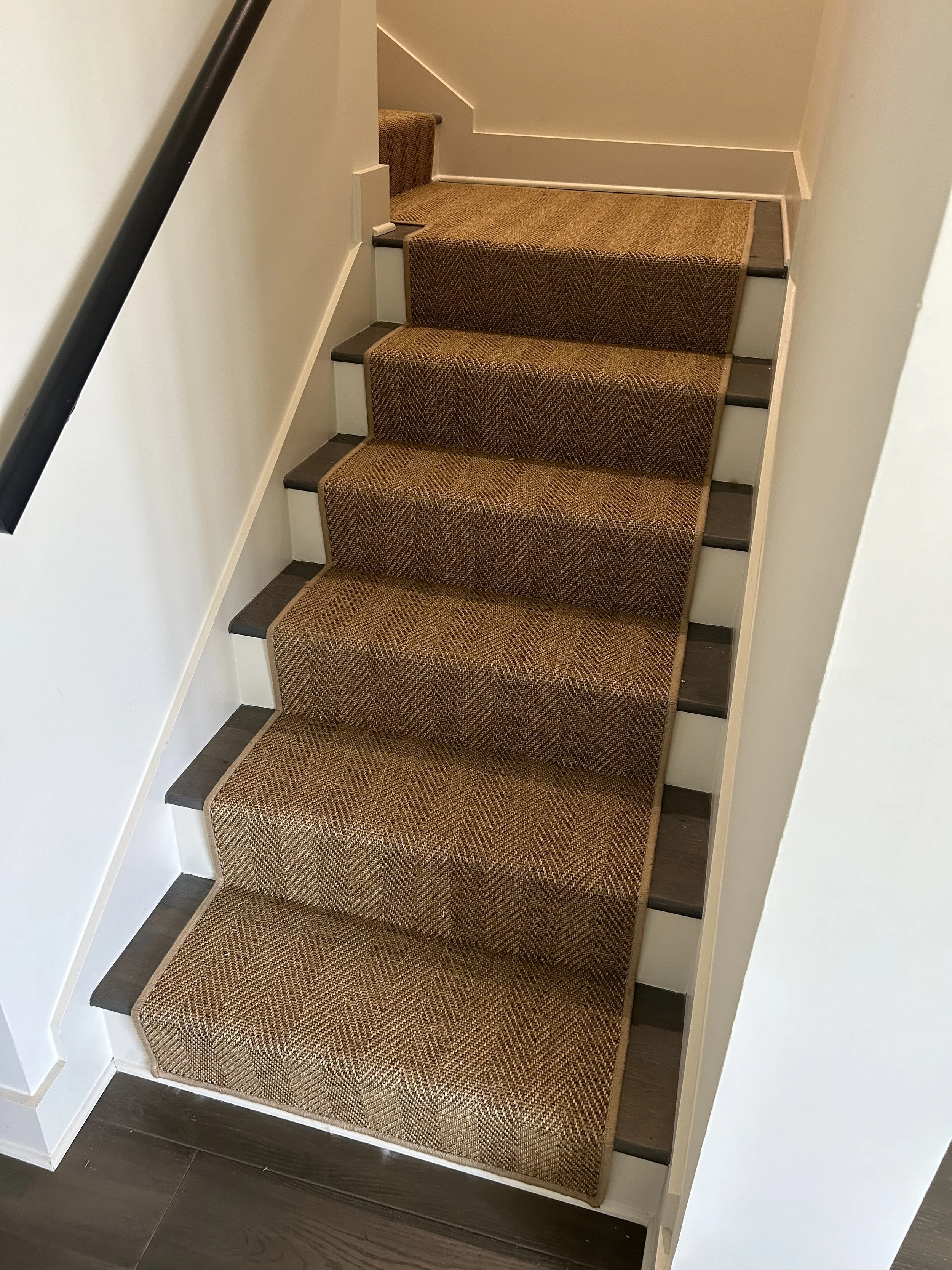 Carpeted staircase with five steps, each with a brown patterned runner, black edge trim, and white risers, adjacent to white walls and a black handrail on the left.