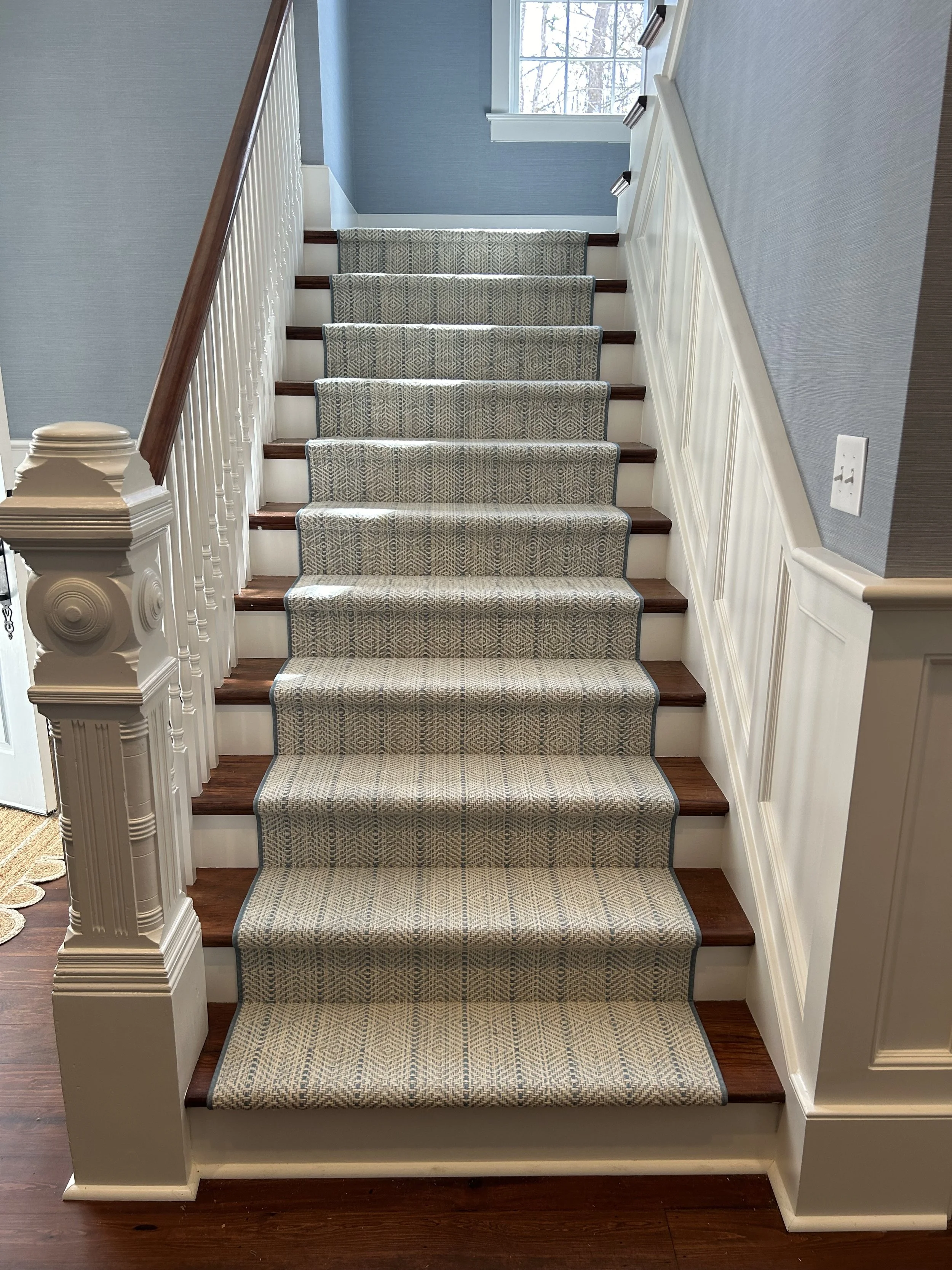 A staircase with wooden steps and a patterned carpet runner, white trim, a balustrade on the left side with decorative white balusters and a wooden handrail, and a window at the top letting in natural light.