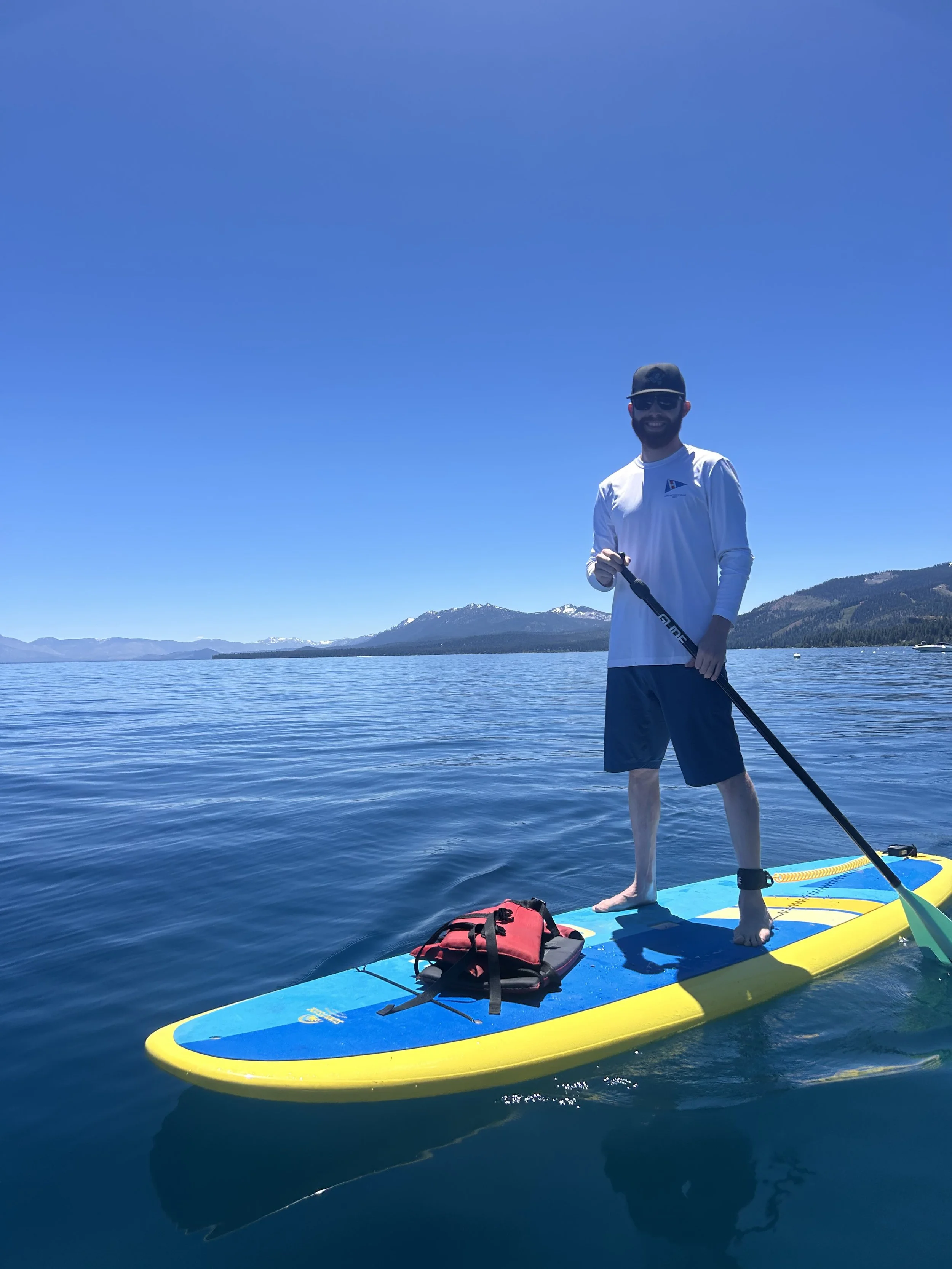 A man with sunglasses, a beard, and wearing a white long-sleeve shirt, shorts, and a cap stands on a paddleboard on a calm lake. The man is smiling and holding a paddle. The paddleboard has a yellow edge and a blue surface. There is a red backpack on the paddleboard, and snow-capped mountains and clear blue sky are visible in the background.