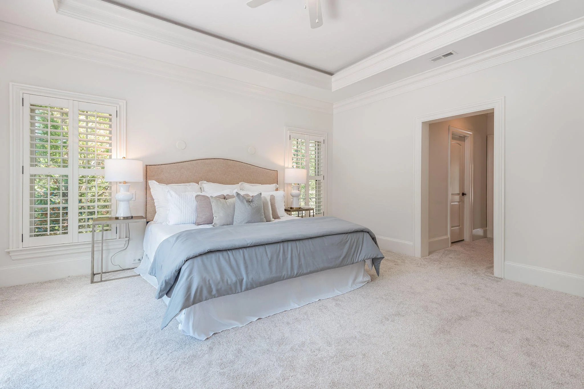 A bedroom with a bed featuring white, gray, and beige pillows, large windows with white shutters, two white lamps on side tables, and a beige carpeted floor.