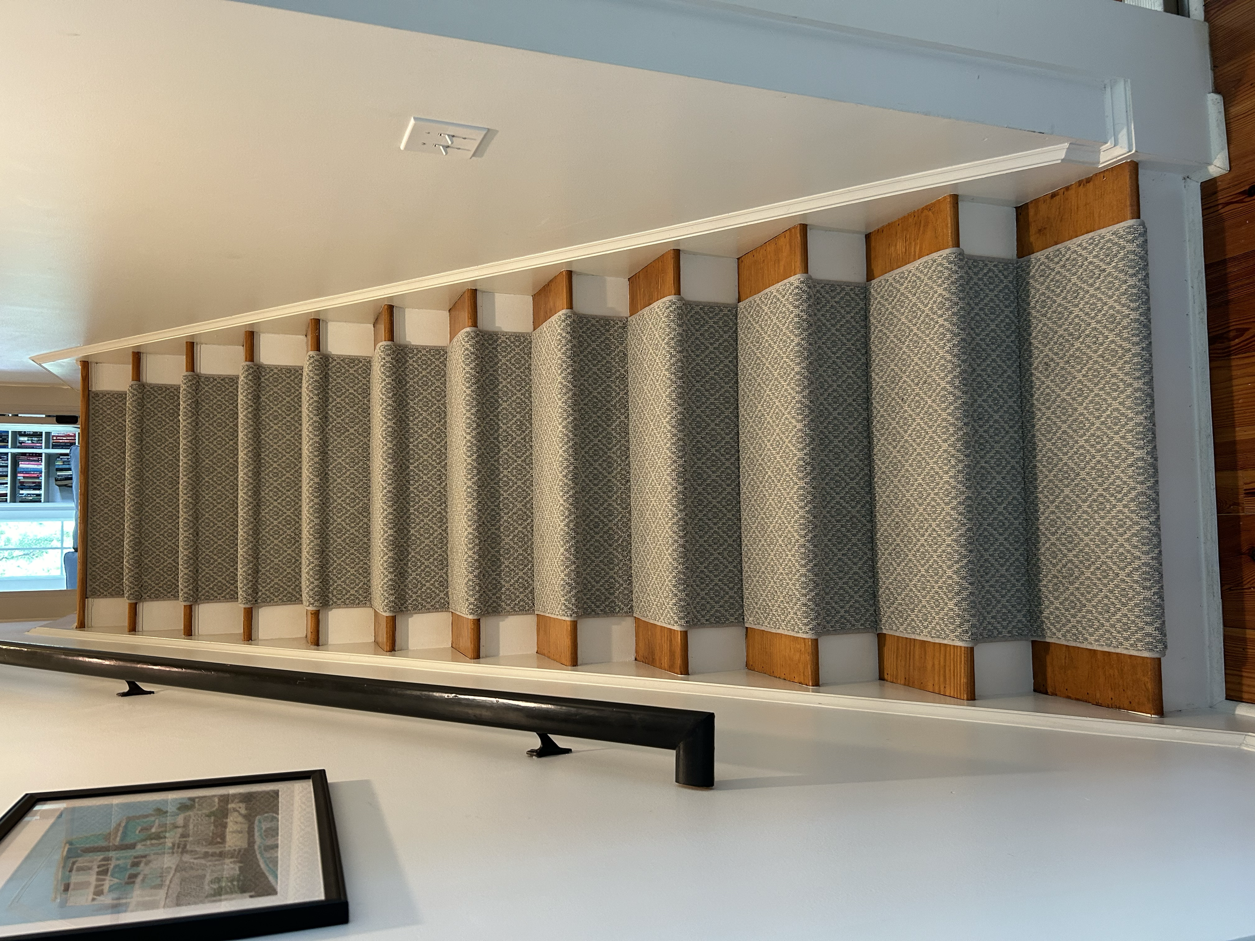 Wooden staircase with gray patterned carpet runner, white walls, a black handrail on the left, a light switch on the right wall, and a framed picture on the left wall. Visible part of the upper floor with a window and a bookshelf in the background.