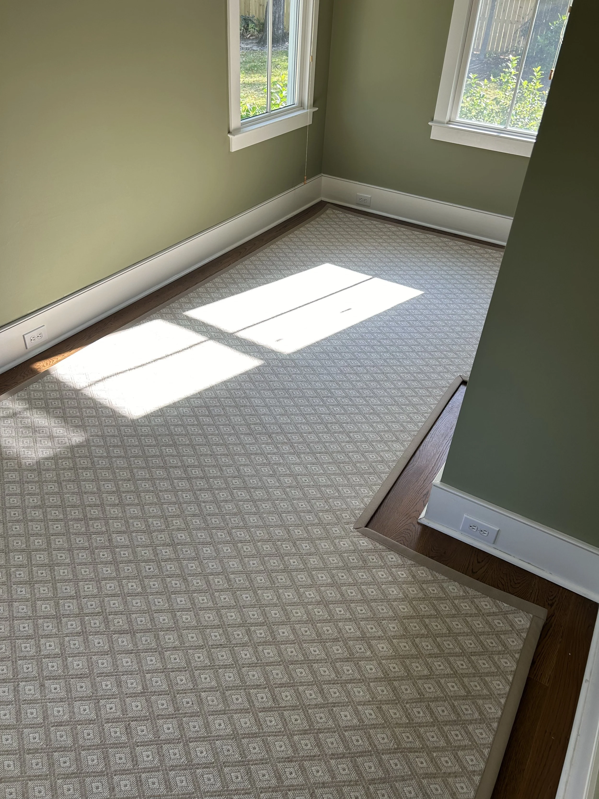 Empty room with beige patterned carpet, green walls, two windows, and electrical outlets.
