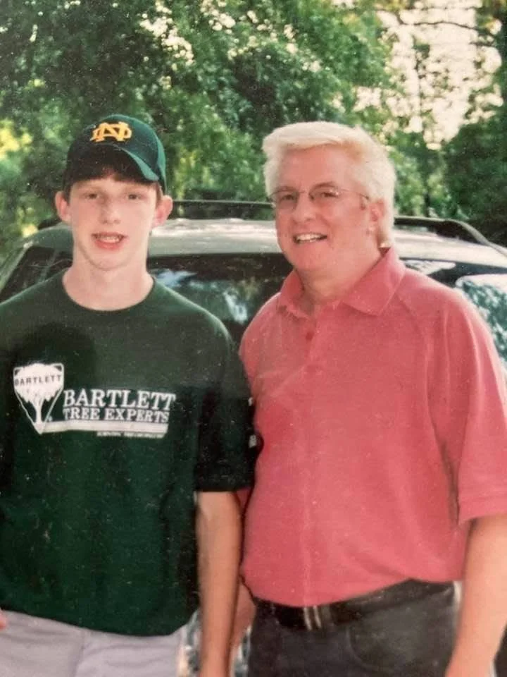Two men standing outdoors, smiling, with a black vehicle and green trees in the background. The younger man on the left is wearing a dark green T-shirt with 'Bartlett Tree Experts' printed on it and a green and yellow baseball cap. The older man on the right has white hair, glasses, and is wearing a pink button-down shirt.