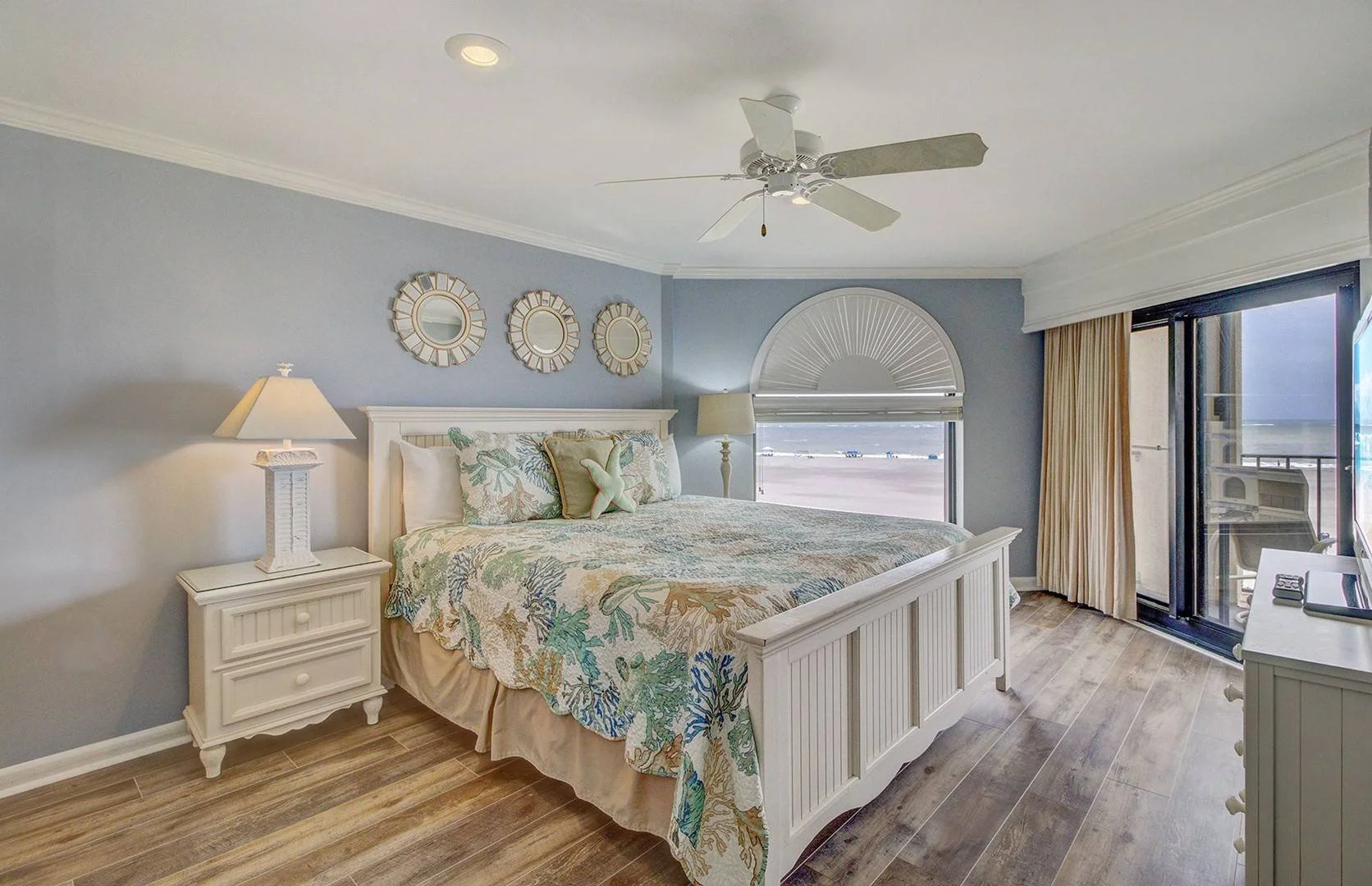 A beachside bedroom with a white wooden bed, tropical-themed bedding, and ocean view through a large window and balcony door.