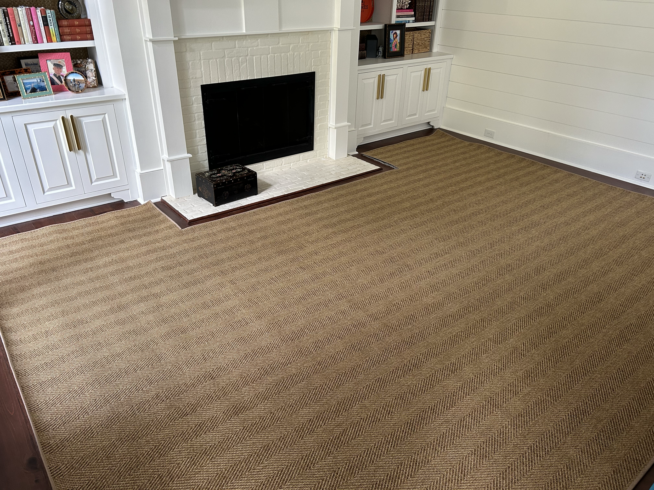 Living room with white built-in shelves, a white fireplace, and a tan woven rug. The shelves contain framed photos, books, and decorative items. The room has white horizontal wall panels and outlets near the floor.