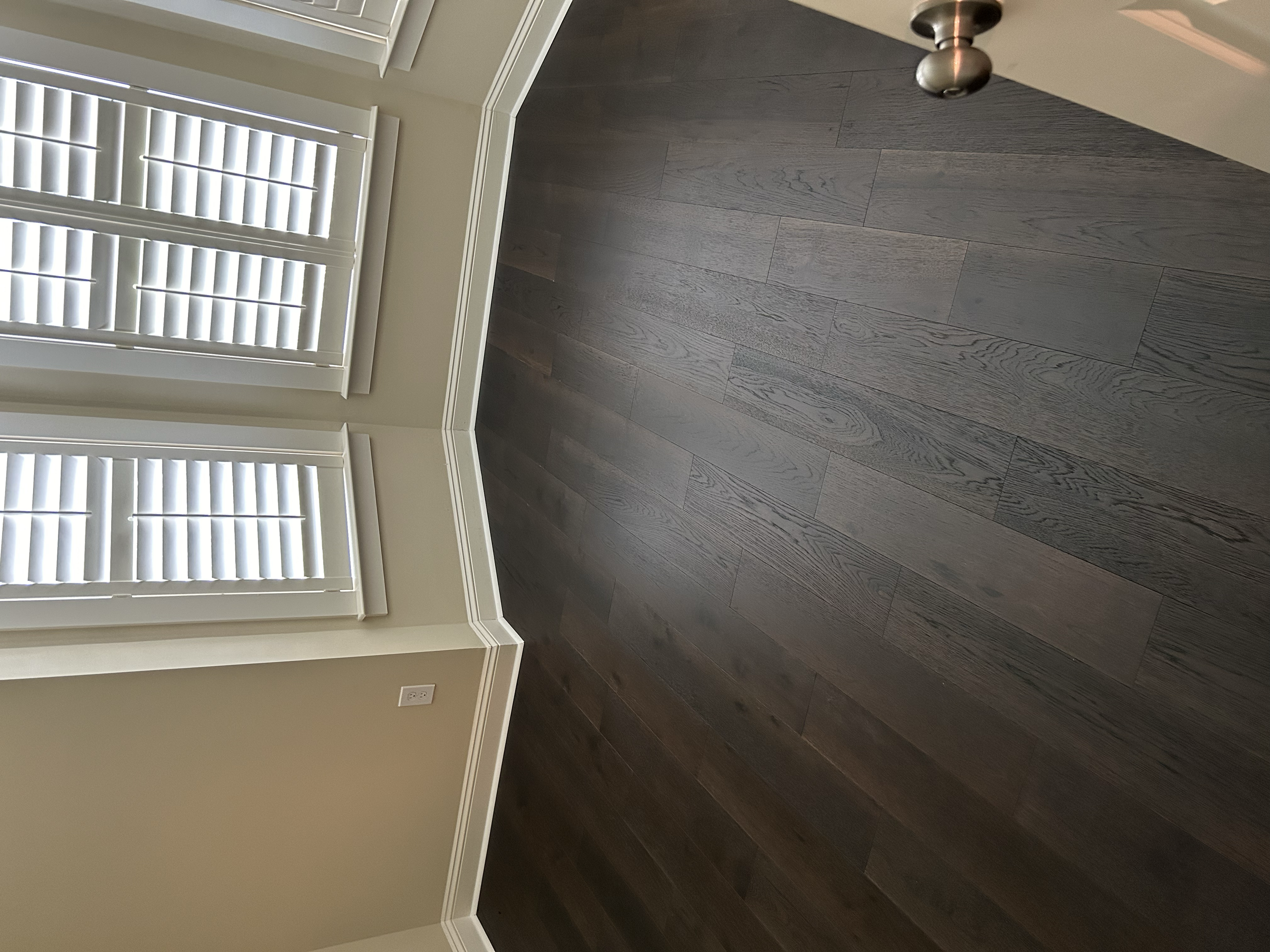 Interior of a room with dark hardwood flooring, beige walls, and white window shutters covering two windows.