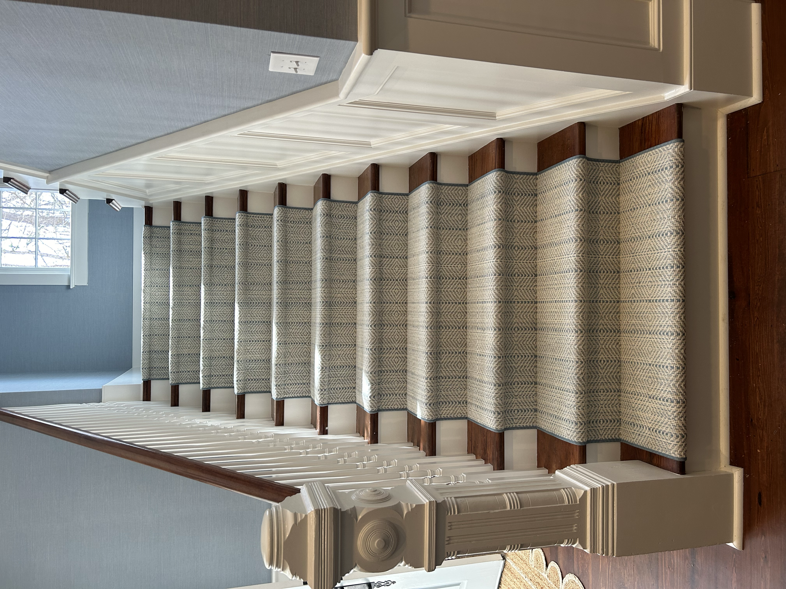 Interior view of a staircase with wooden steps, a patterned carpet runner, white painted walls, decorative trim, and a window letting in natural light.
