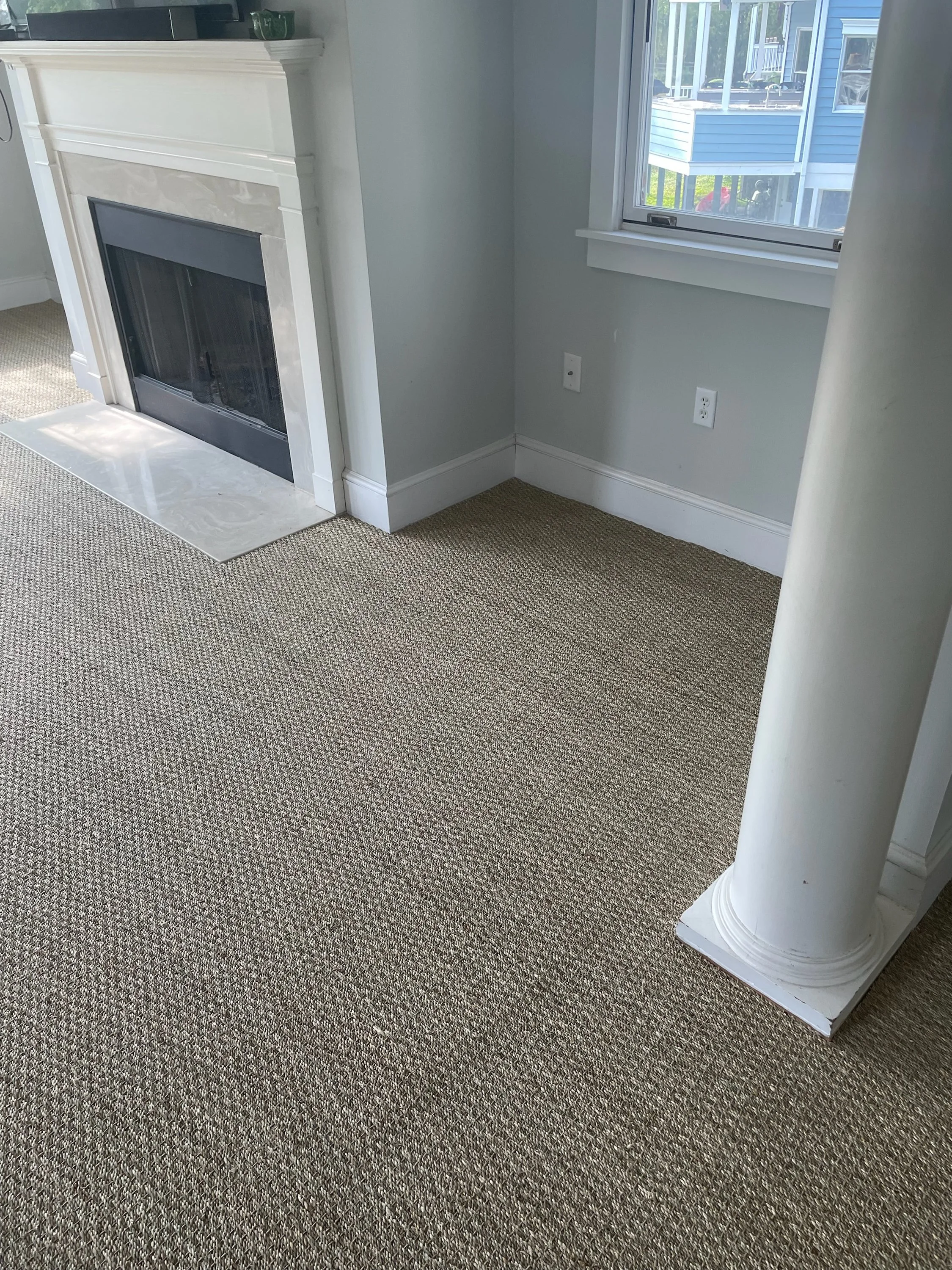 Corner of a room with a fireplace, a window showing a house outside, and a beige carpet.