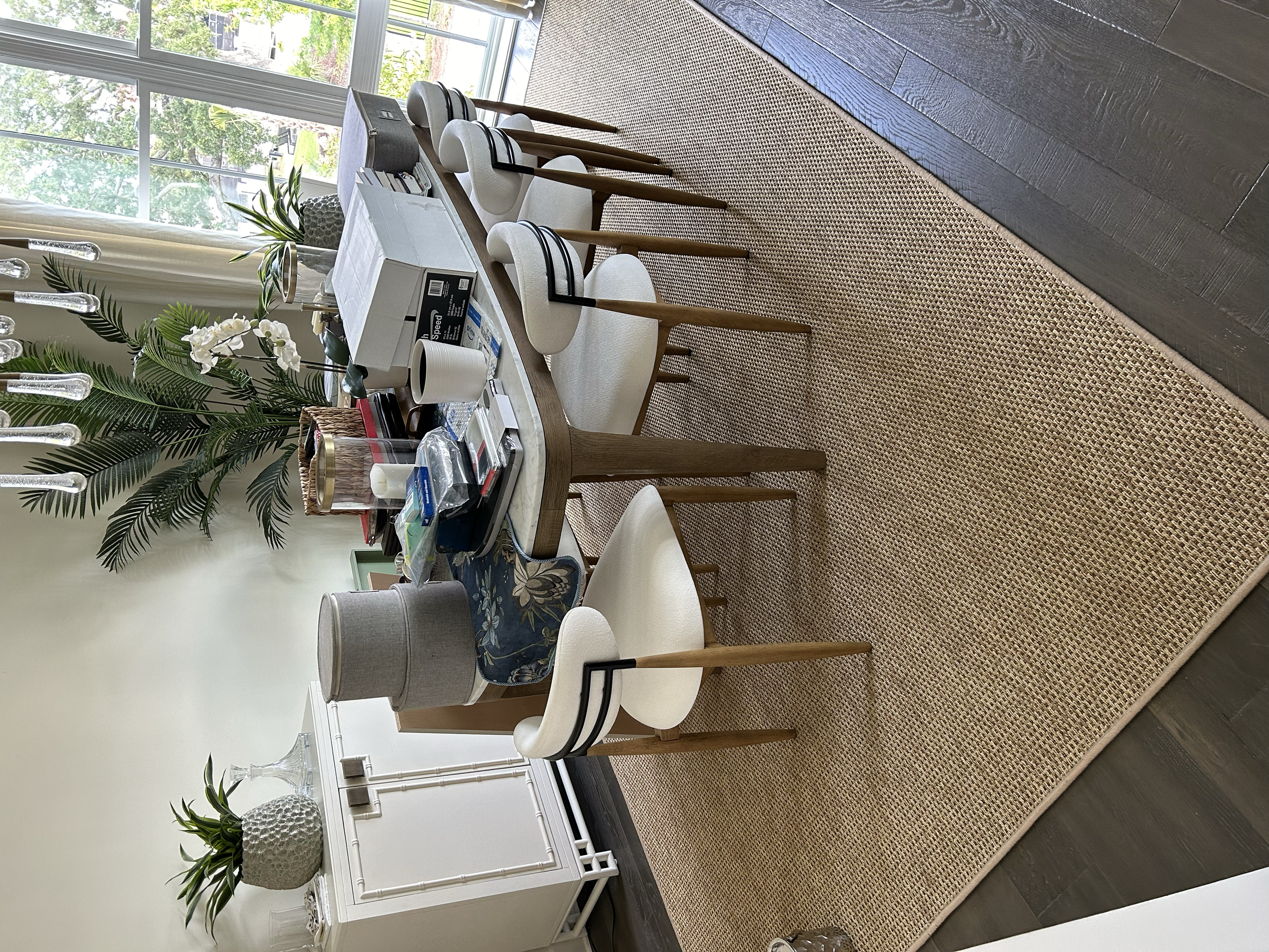 A rectangular dining table with six white chairs, a woven rug underneath, and various decorative items on the table including boxes, a candle, and plants. In the background, there is a white sideboard with potted plants and glass decor, and large win