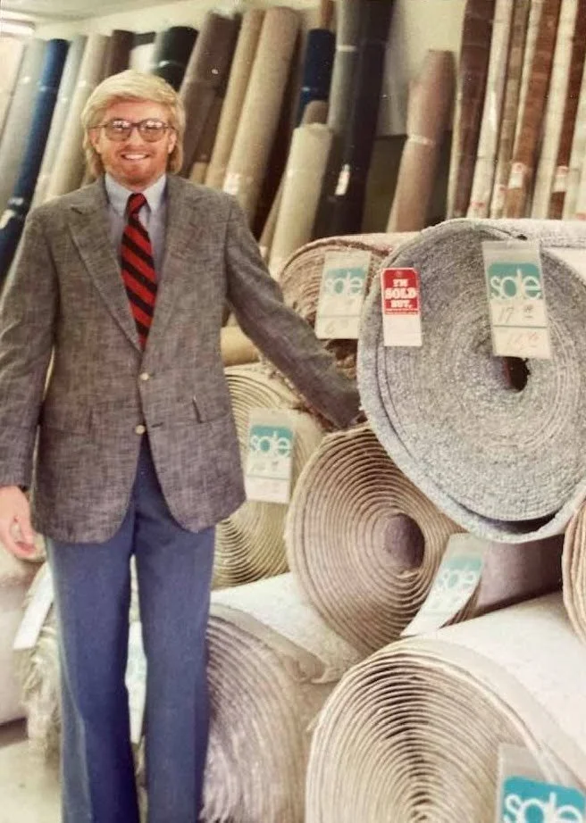 A man with glasses, blonde hair, and a beard wearing a plaid blazer, light blue shirt, and red striped tie, standing in a store aisle next to large rolls of carpet or fabric, some marked with sale tags.