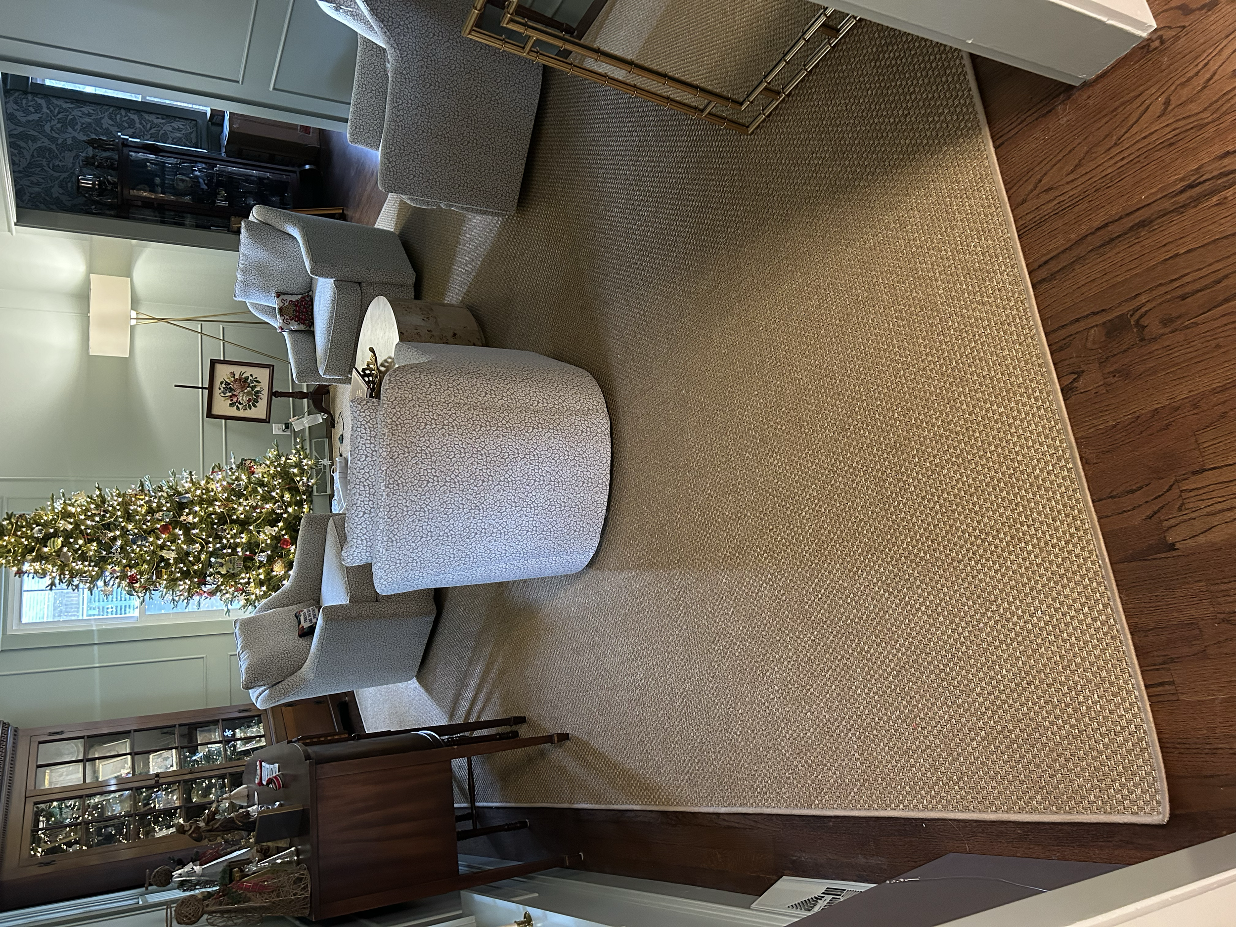 Living room with a decorated Christmas tree, four upholstered chairs, a wooden cabinet, a floor lamp, and a floral artwork on a stand. The room has hardwood floors and a beige area rug.