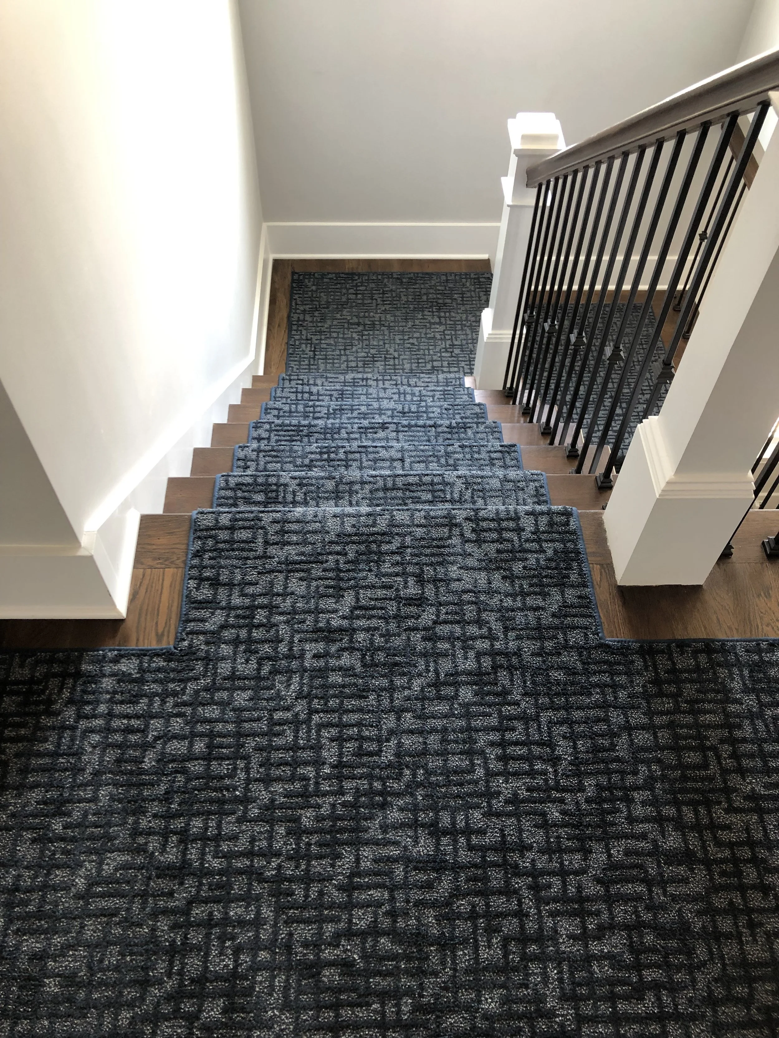 View of a staircase with dark patterned carpet, wooden steps, and black metal balusters, leading down to a small landing with a darker rug, bordered by white walls and a white handrail.