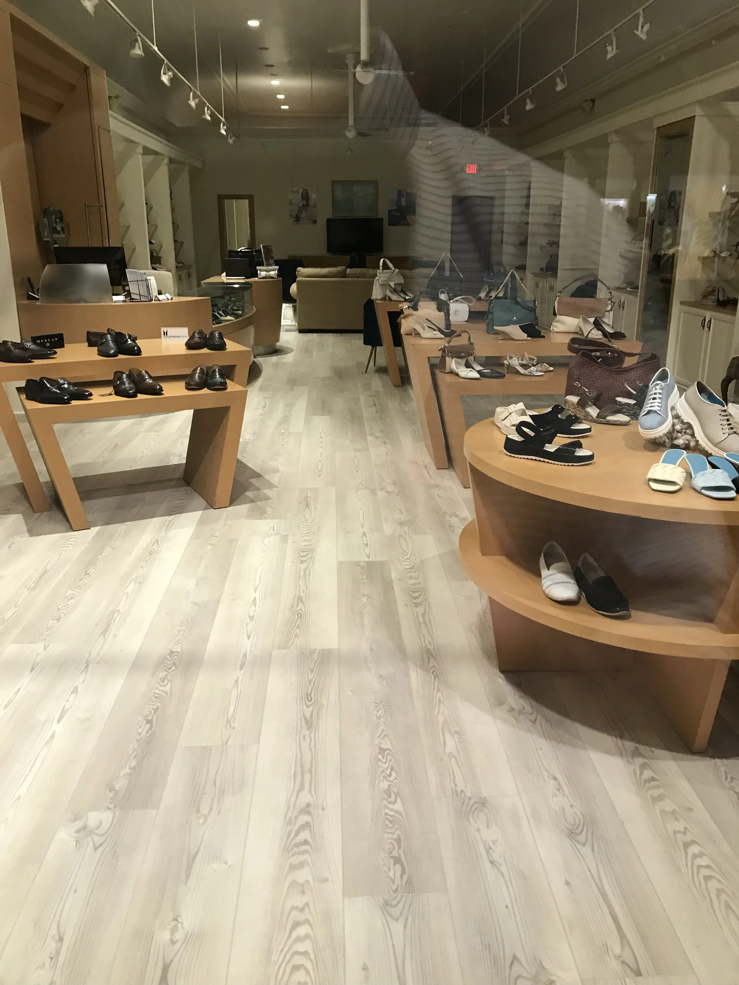 Interior of a shoe store with wooden display tables showing various shoes and bags, with a seating area and television in the background.
