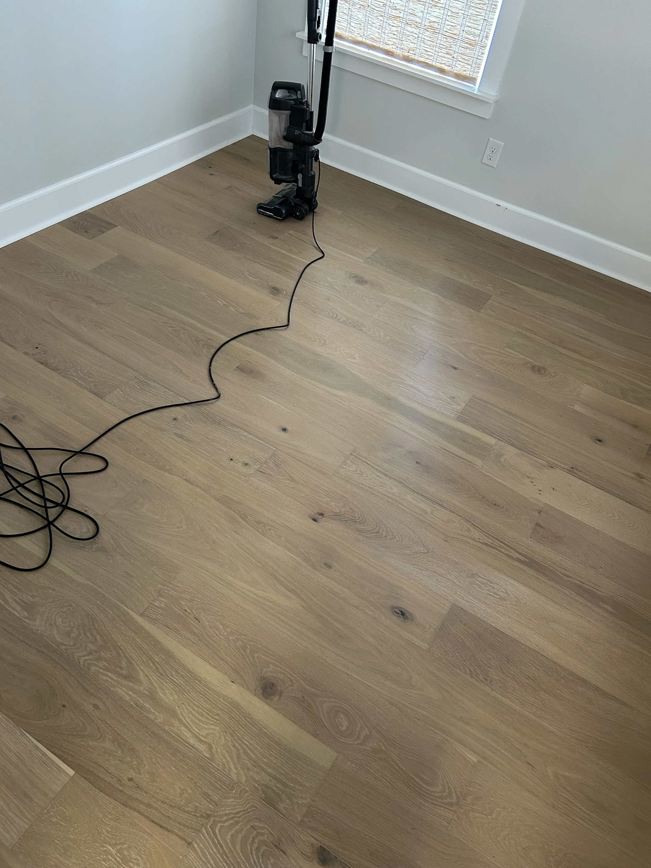 A vacuum cleaner standing on a hardwood floor in a corner of a room near a window with a bamboo shade and an electrical outlet.