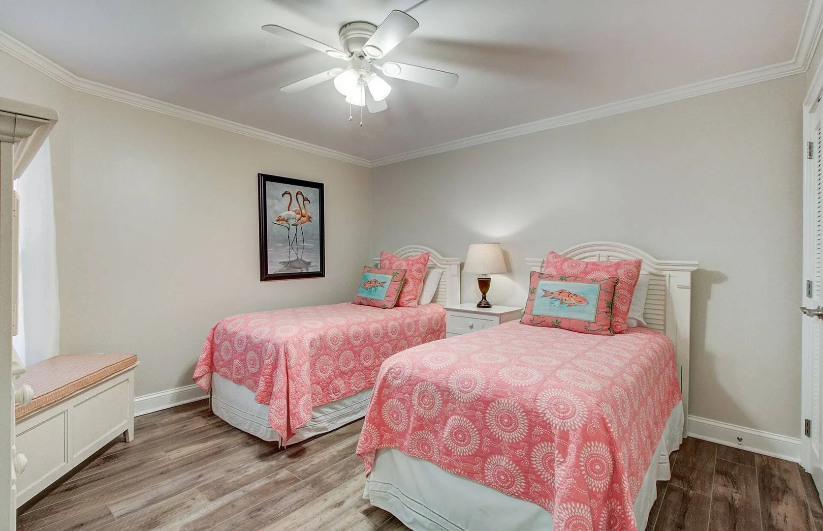 A bedroom with two twin beds featuring pink patterned bedspreads and decorative pillows with fish designs. There is a white nightstand with a lamp between the beds, a ceiling fan with lights, a fish-themed painting on the wall, a window with white cu