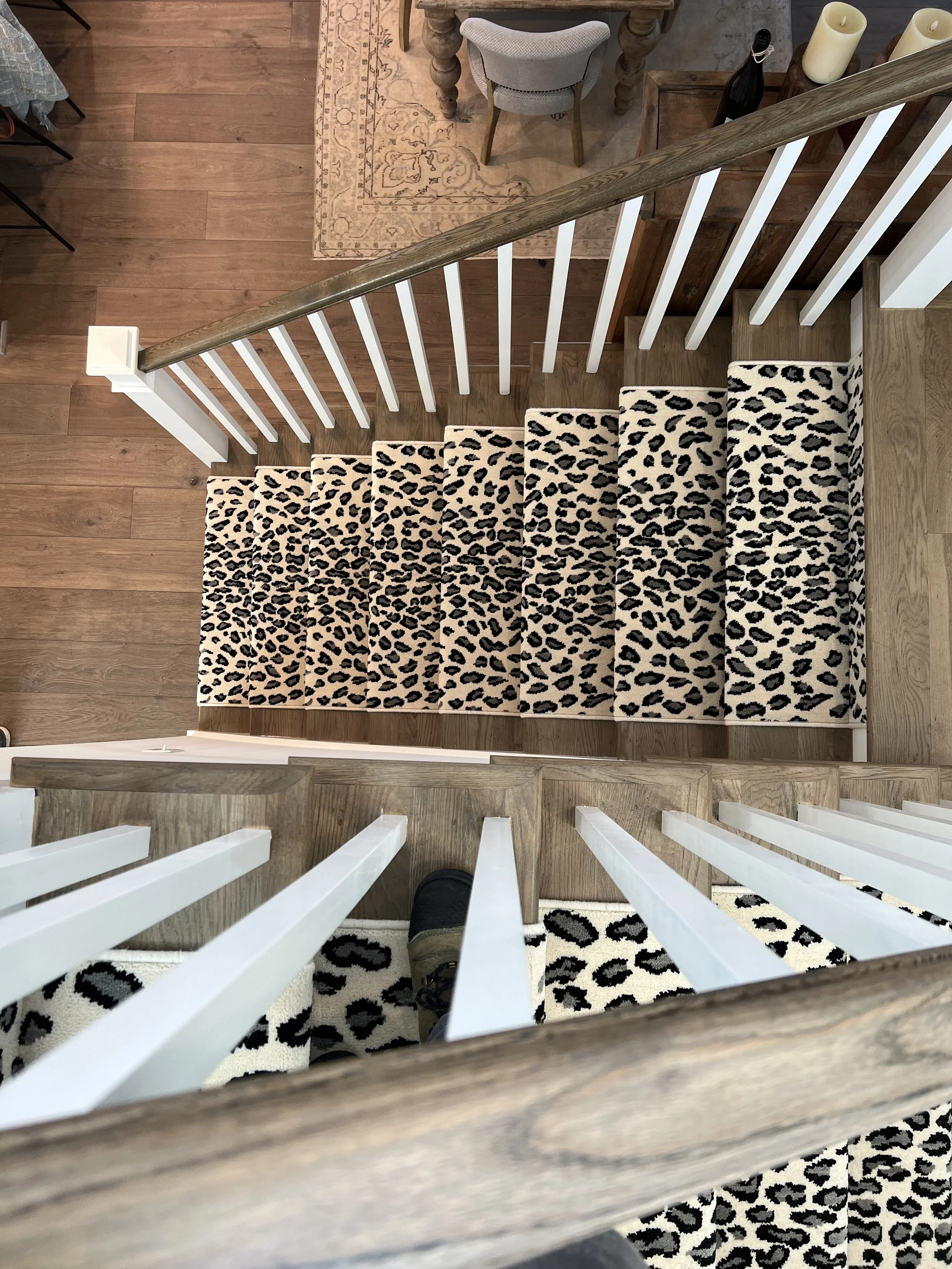 View looking down at the bottom of a staircase with a leopard print rug at the foot of the stairs, a wooden handrail, and white balusters. Part of a room with hardwood flooring, a beige patterned rug, a desk, a chair, candles, and decorative bottles 