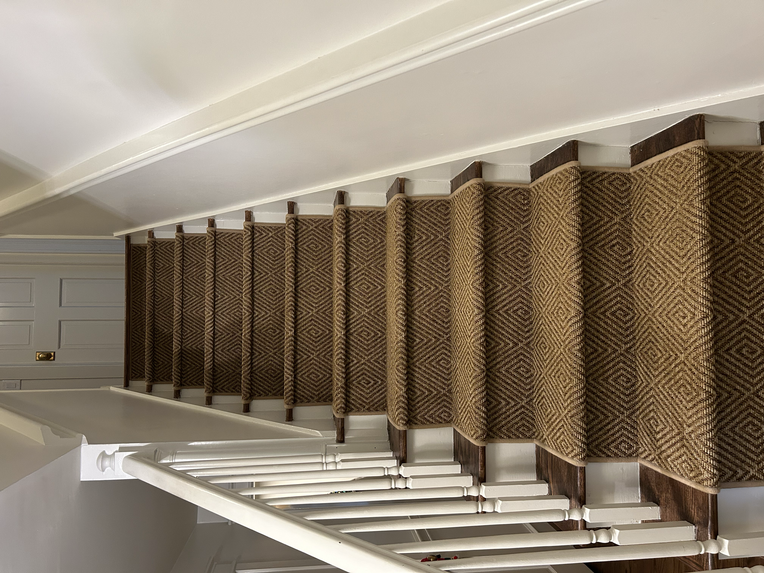 Indoor staircase with carpeted steps, white walls, a white railing on the left, and a closed door at the top of the stairs.
