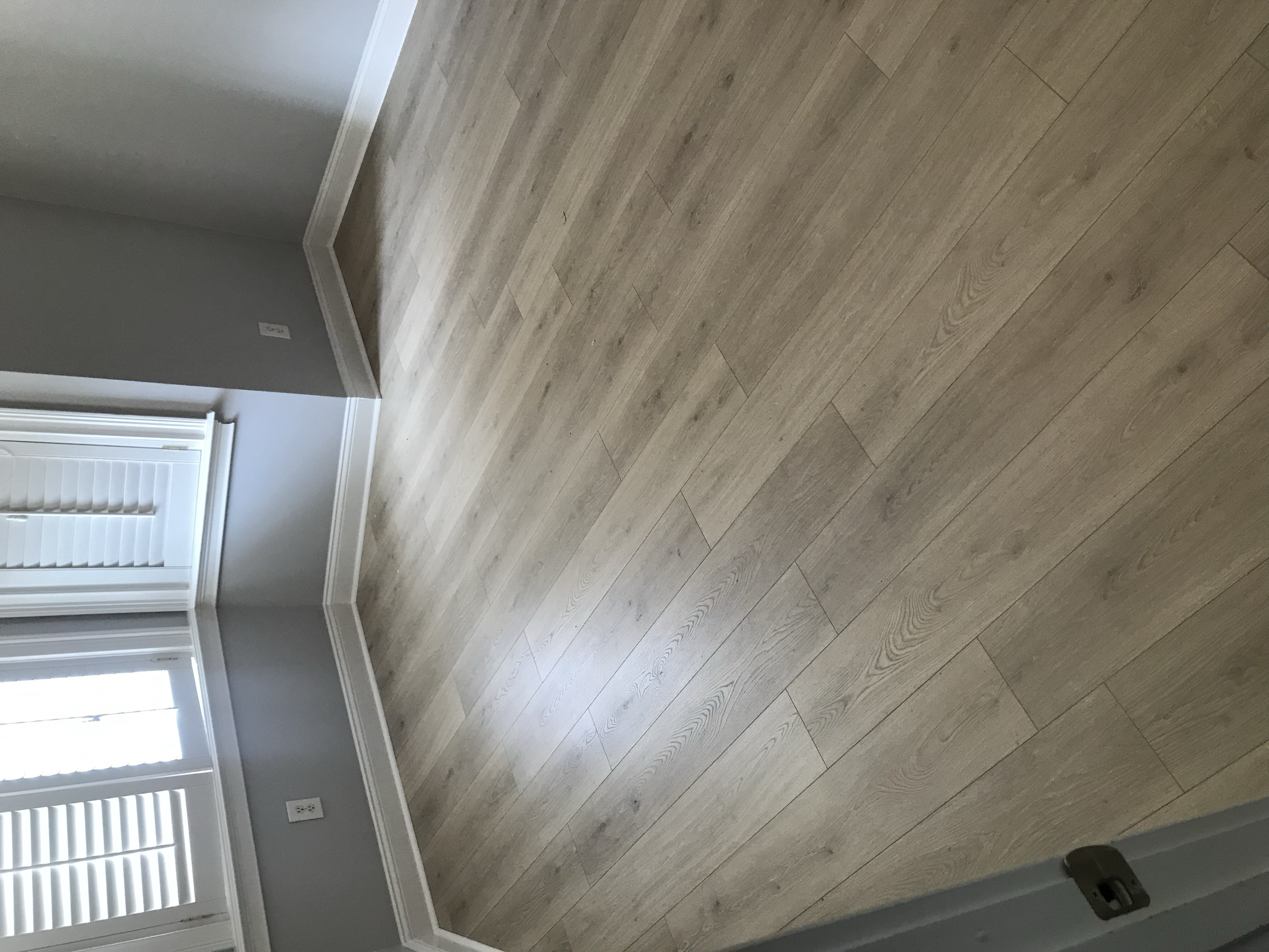 An empty room with light wood flooring, gray walls, white baseboards, and white window shutters.