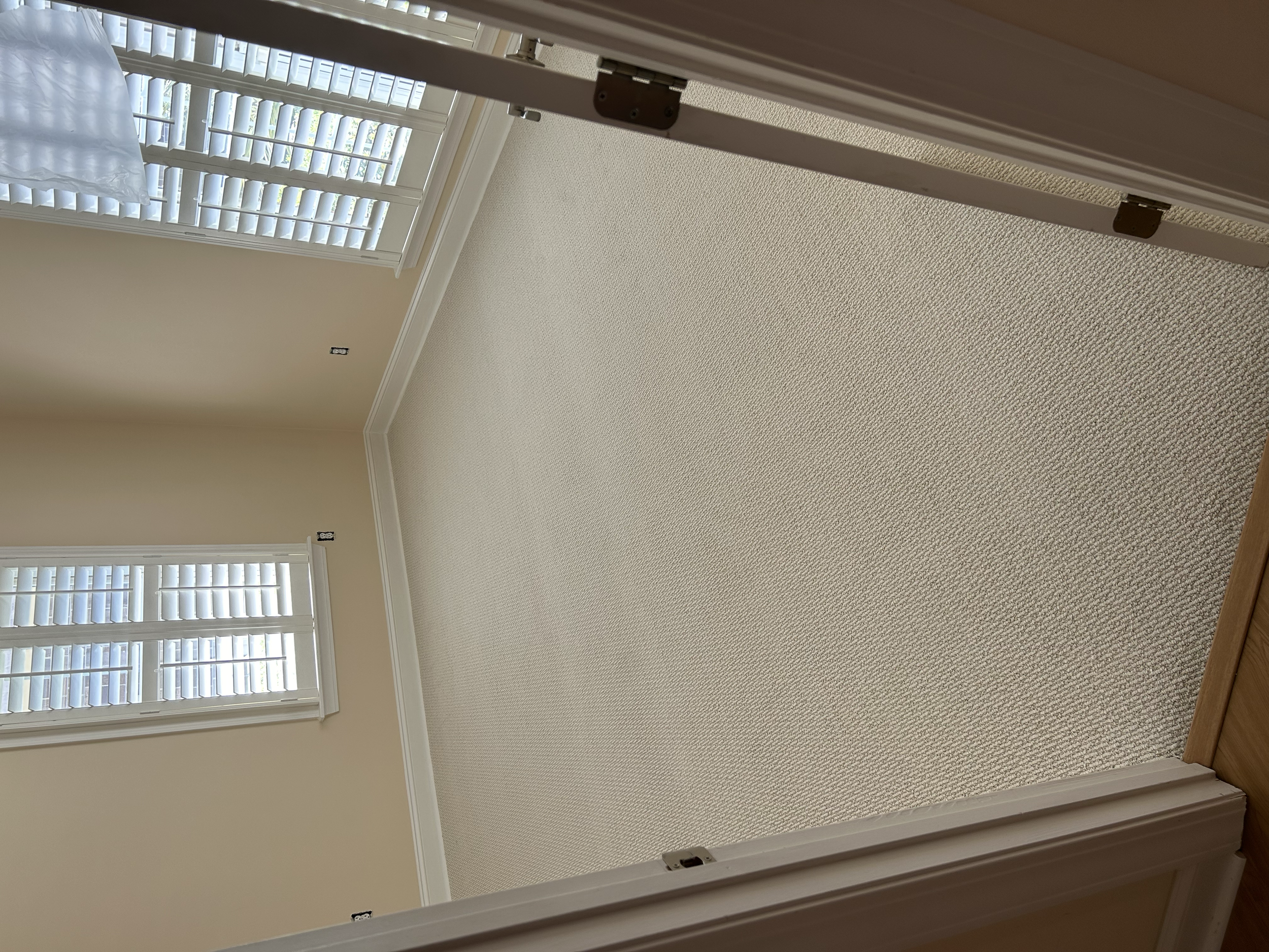 Empty room with beige walls, white shutters on windows, and beige carpeted flooring.