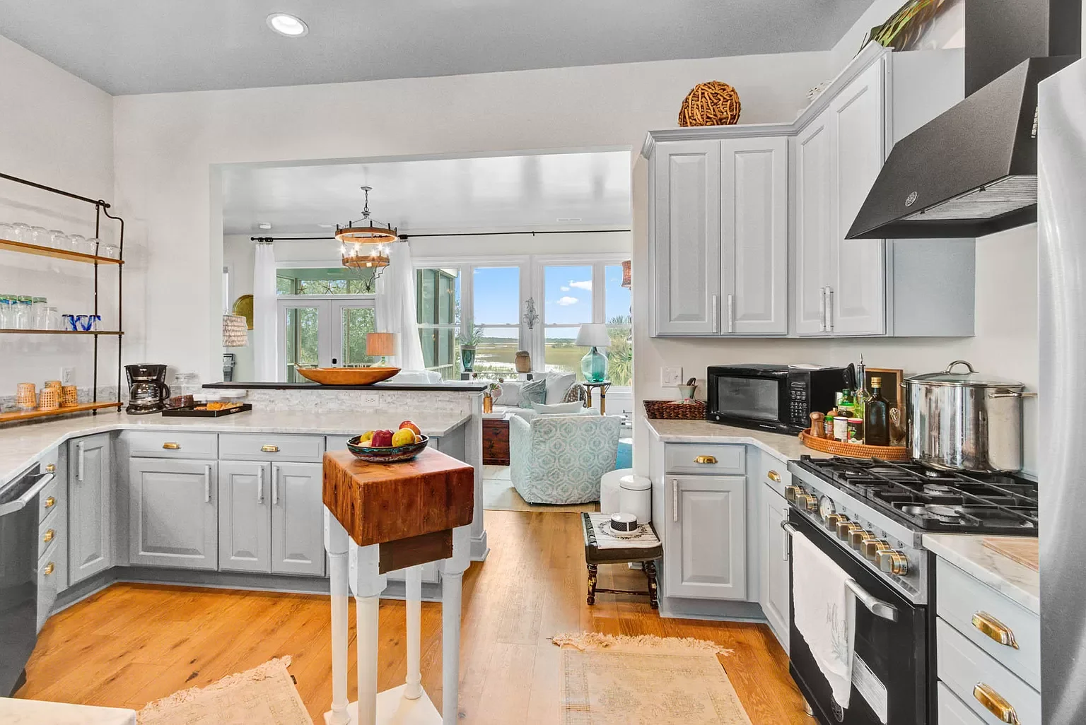Kitchen with white cabinets, black and silver appliances, a small wooden table with a bowl of apples, open shelves with glasses, and a view into a bright living room with a view of outdoors.
