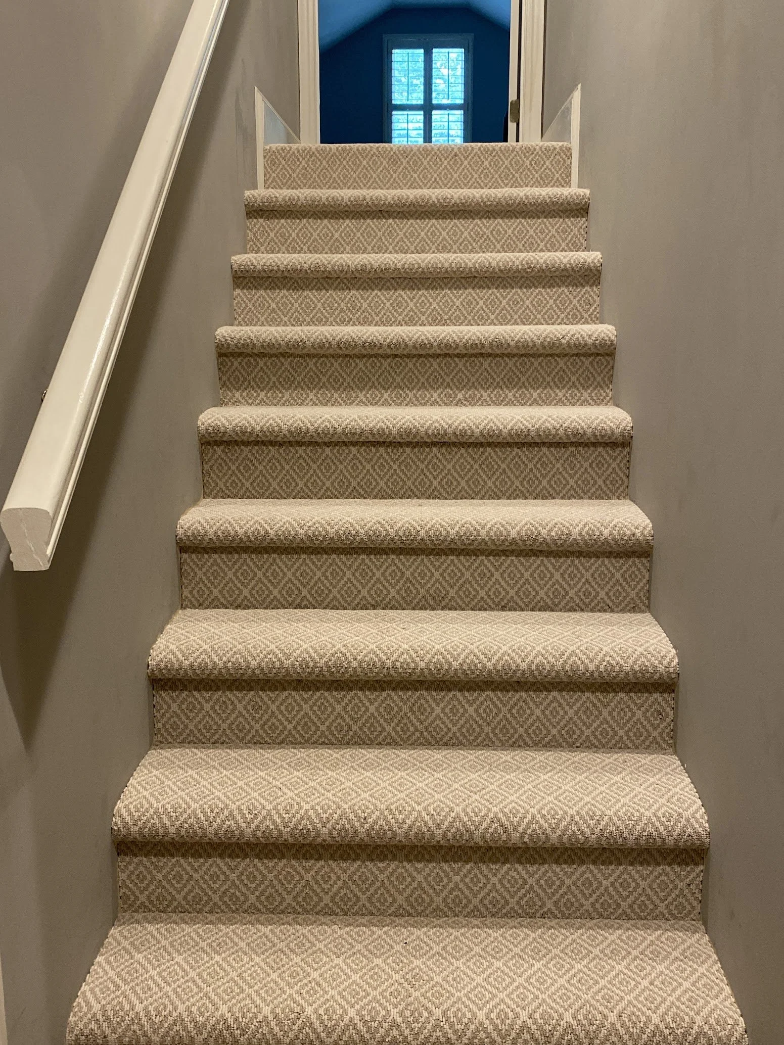 Indoor staircase with beige patterned carpet, gray walls, white trim, leading up to a blue room with a window.