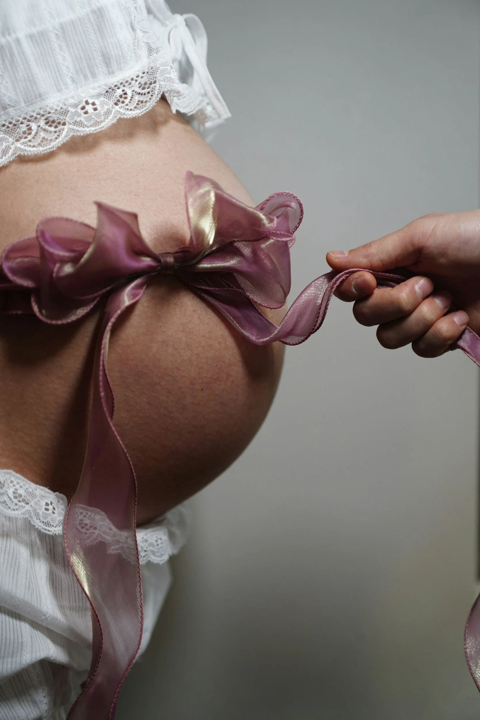 Close-up of a pregnant woman's belly with a large pink ribbon tied in a bow around her waist, wearing a white lace top or dress.