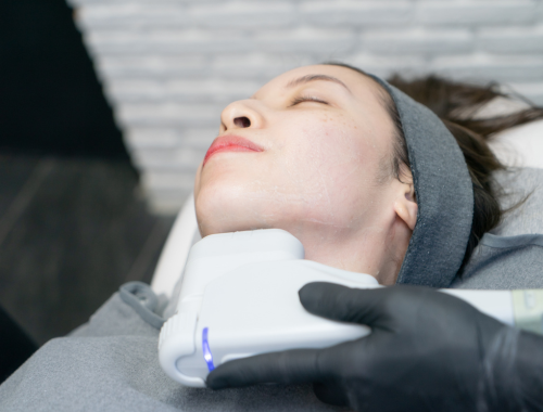 A young woman lying on a medical examination table with her eyes closed, receiving a painless laser or IPL skin treatment on her jawline from a healthcare professional wearing black gloves.