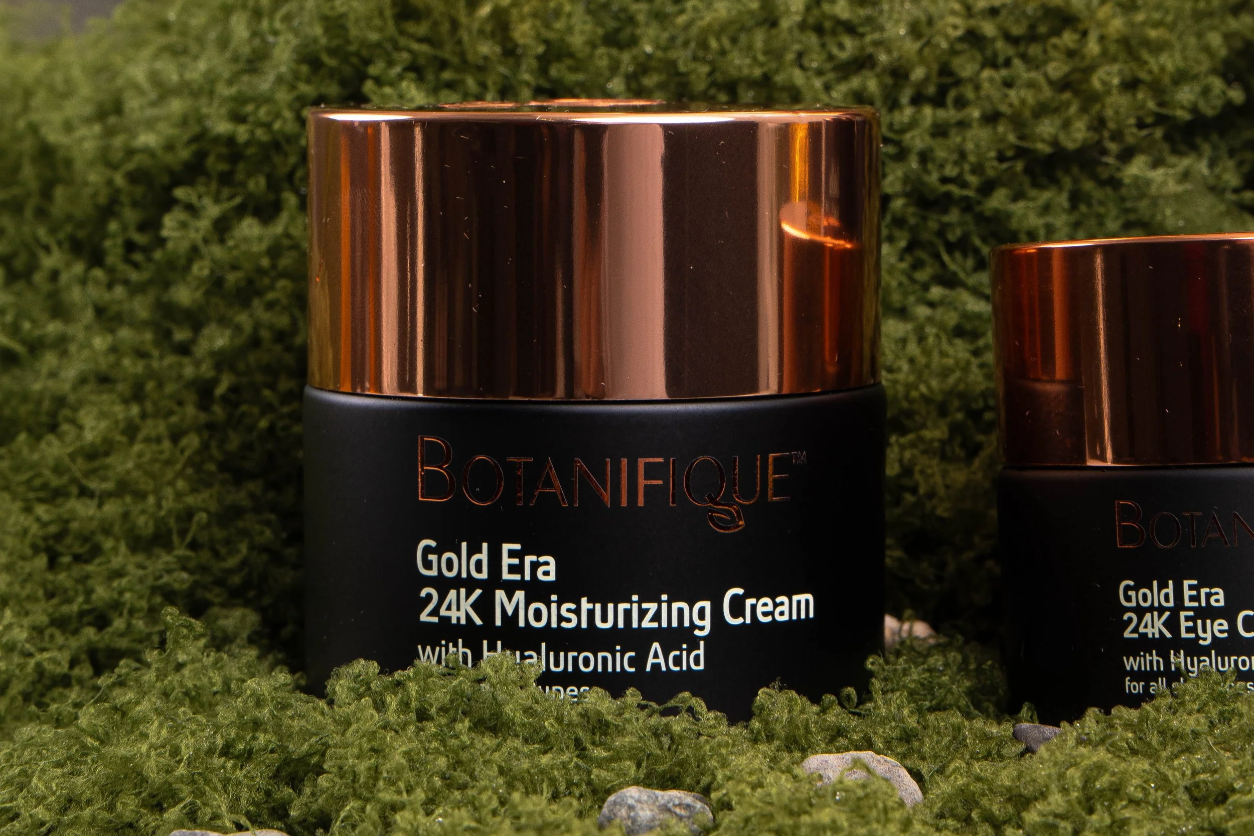 Close-up of the Botanifique Gold Era 24K Moisturizing Cream in black and rose gold packaging, displayed among green moss with the matching eye cream partially visible.