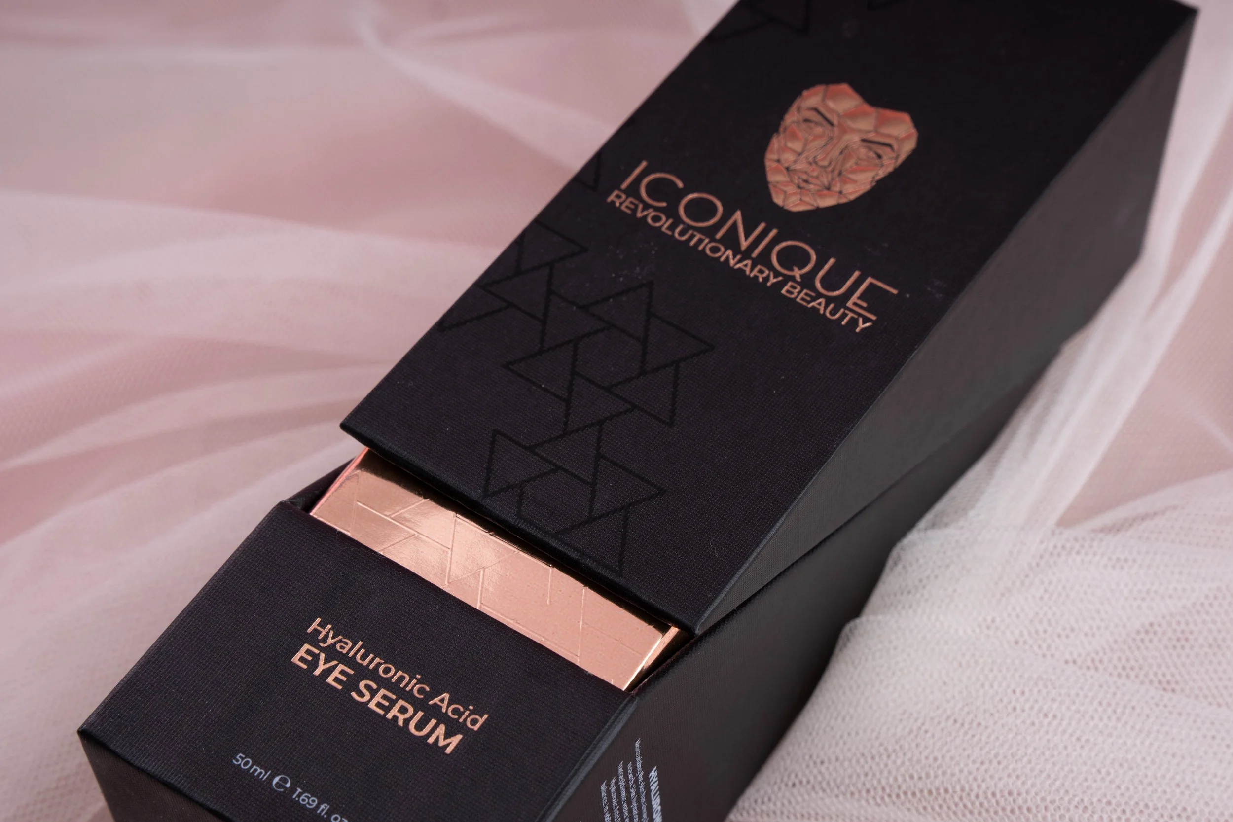 Close-up of the ICONIQUE Hyaluronic Acid Eye Serum packaging featuring elegant black boxes with rose gold logo and product details displayed on soft white fabric.