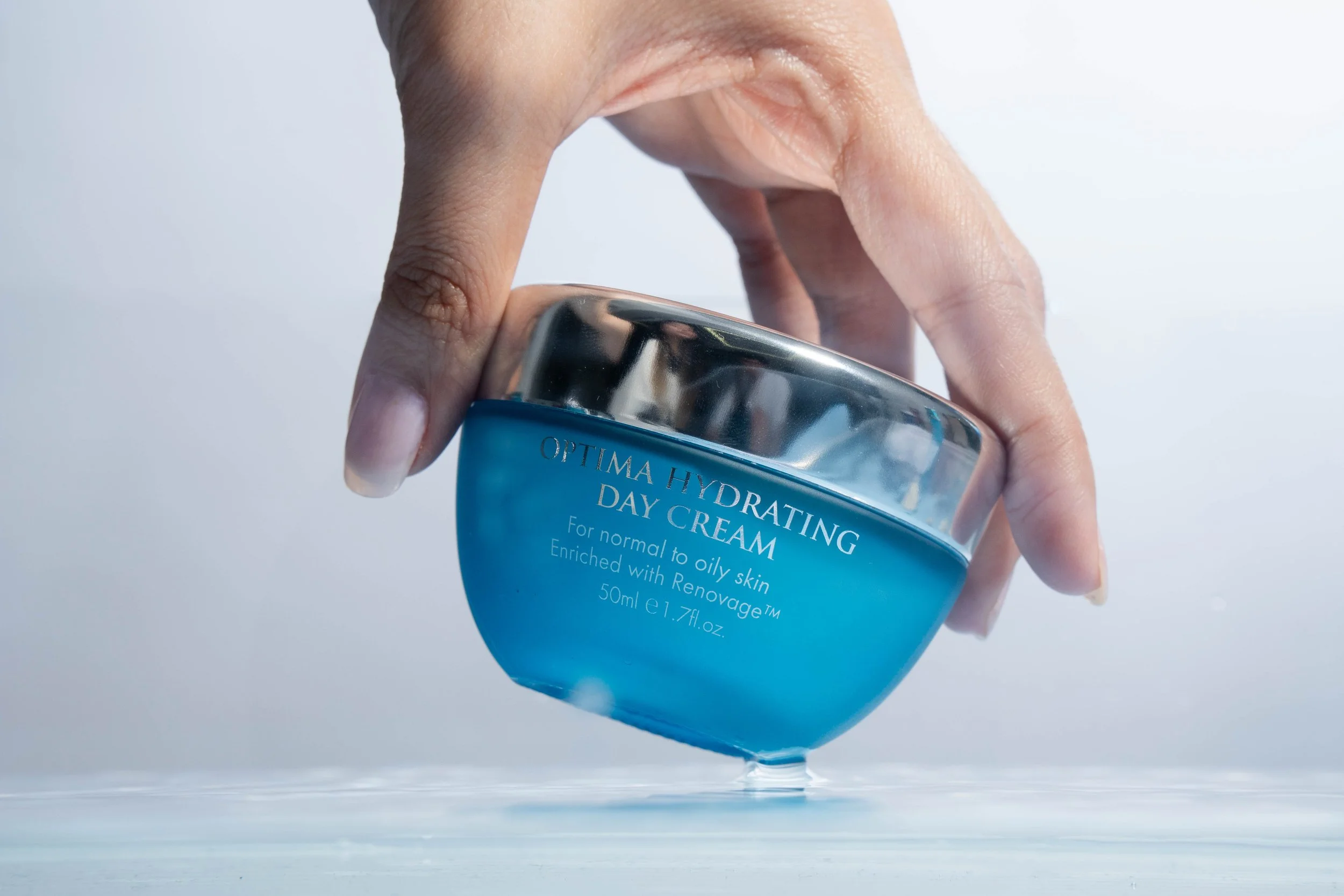 Close-up of a hand holding the blue Aqua Mineral Optima Hydrating Day Cream jar with a reflective silver lid above a glossy surface.