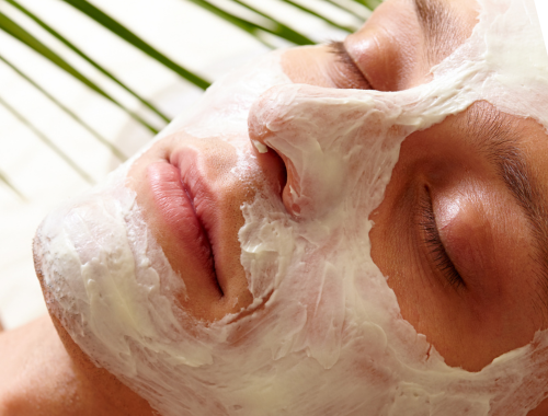 A woman receiving a facial treatment with white cream mask applied to her face, lying down with eyes closed.