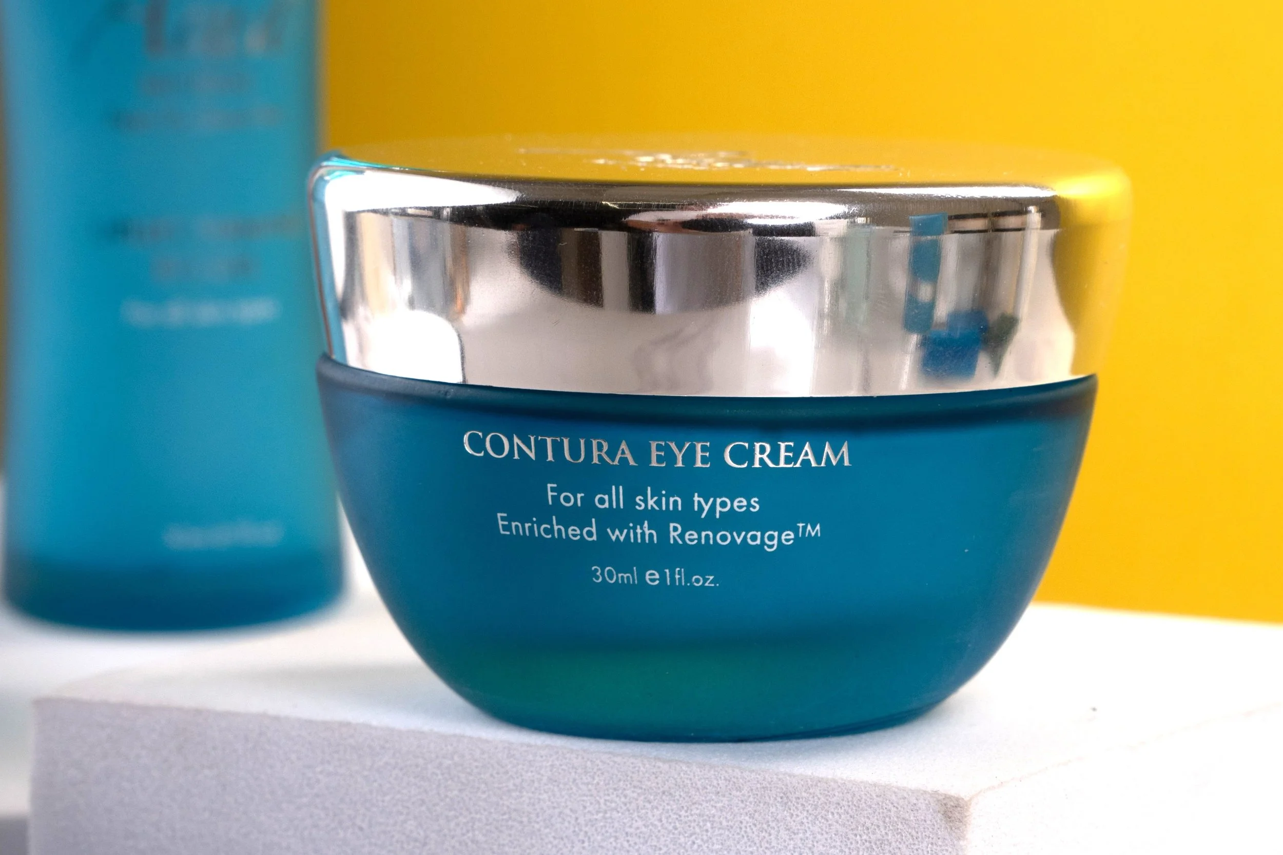 Close-up of the Aqua Mineral Contura Eye Cream in a blue jar with a reflective silver lid, displayed on a white pedestal with a blue and yellow backdrop.