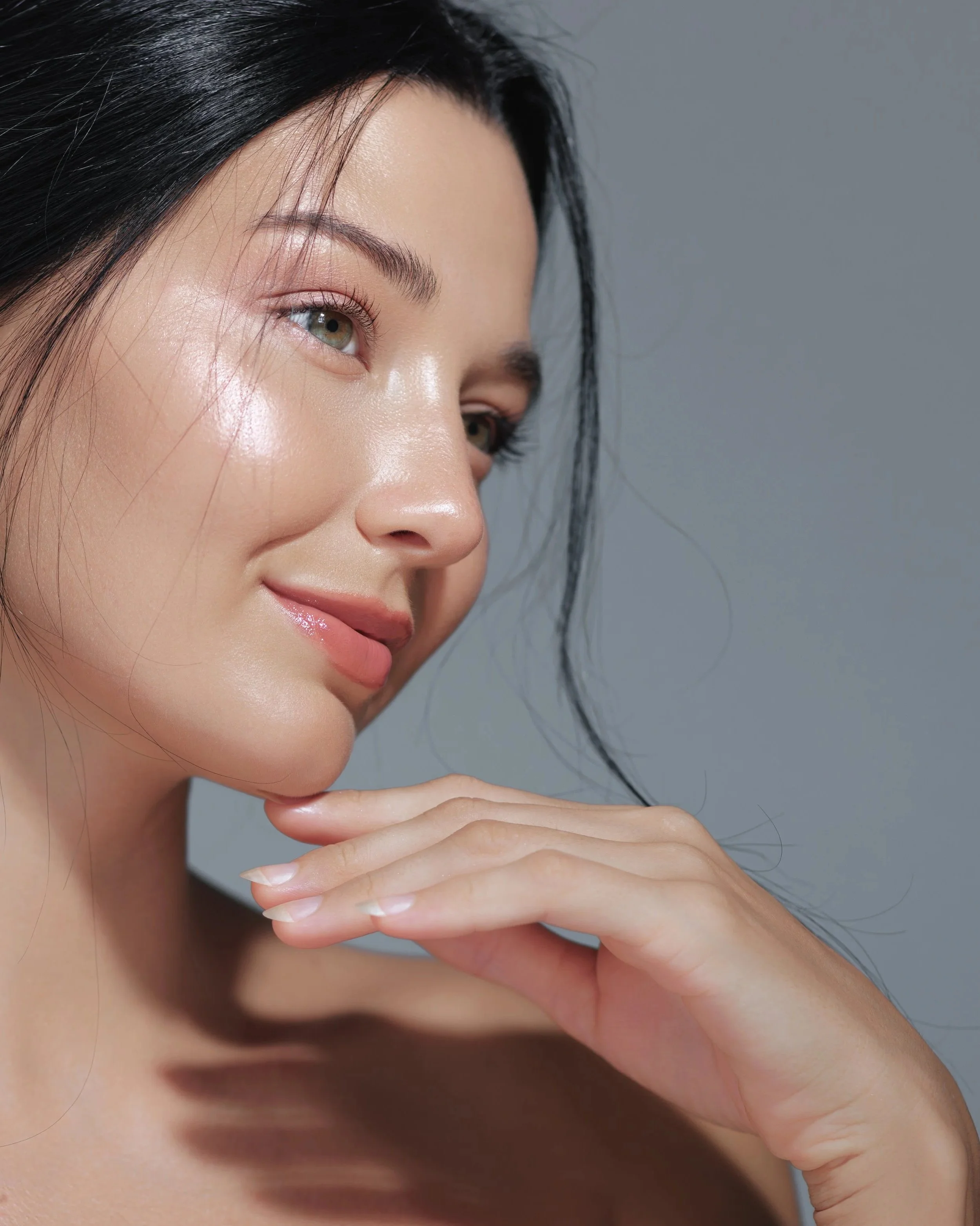 Close-up of a woman with black hair, glowing skin, and natural makeup, gently touching her face against a gray background.