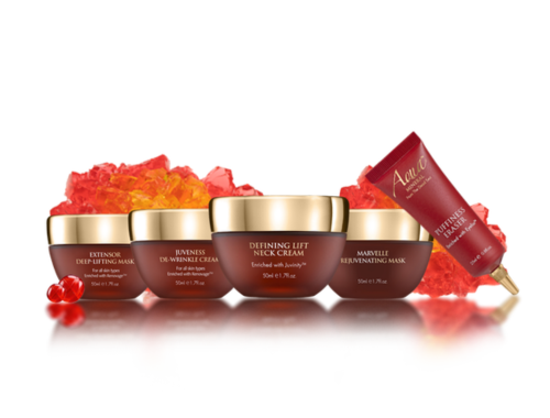 A collection of four skincare jars and a red tube with gold caps, placed on a reflective surface behind red and orange textured elements and small red beads.