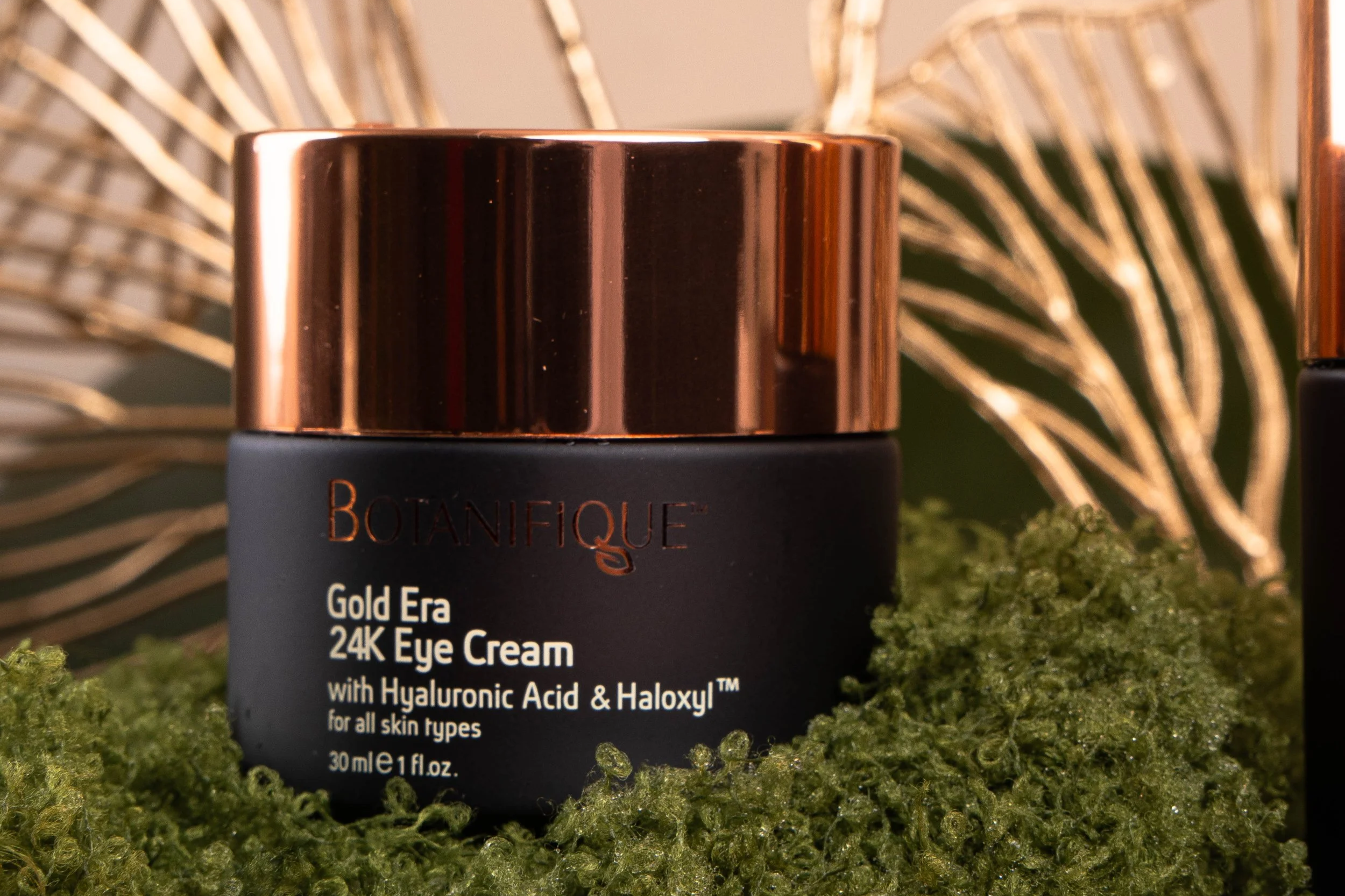 Botanifique Gold Era 24K Eye Cream with Hyaluronic Acid and Haloxyl displayed in black and rose gold packaging among green moss.