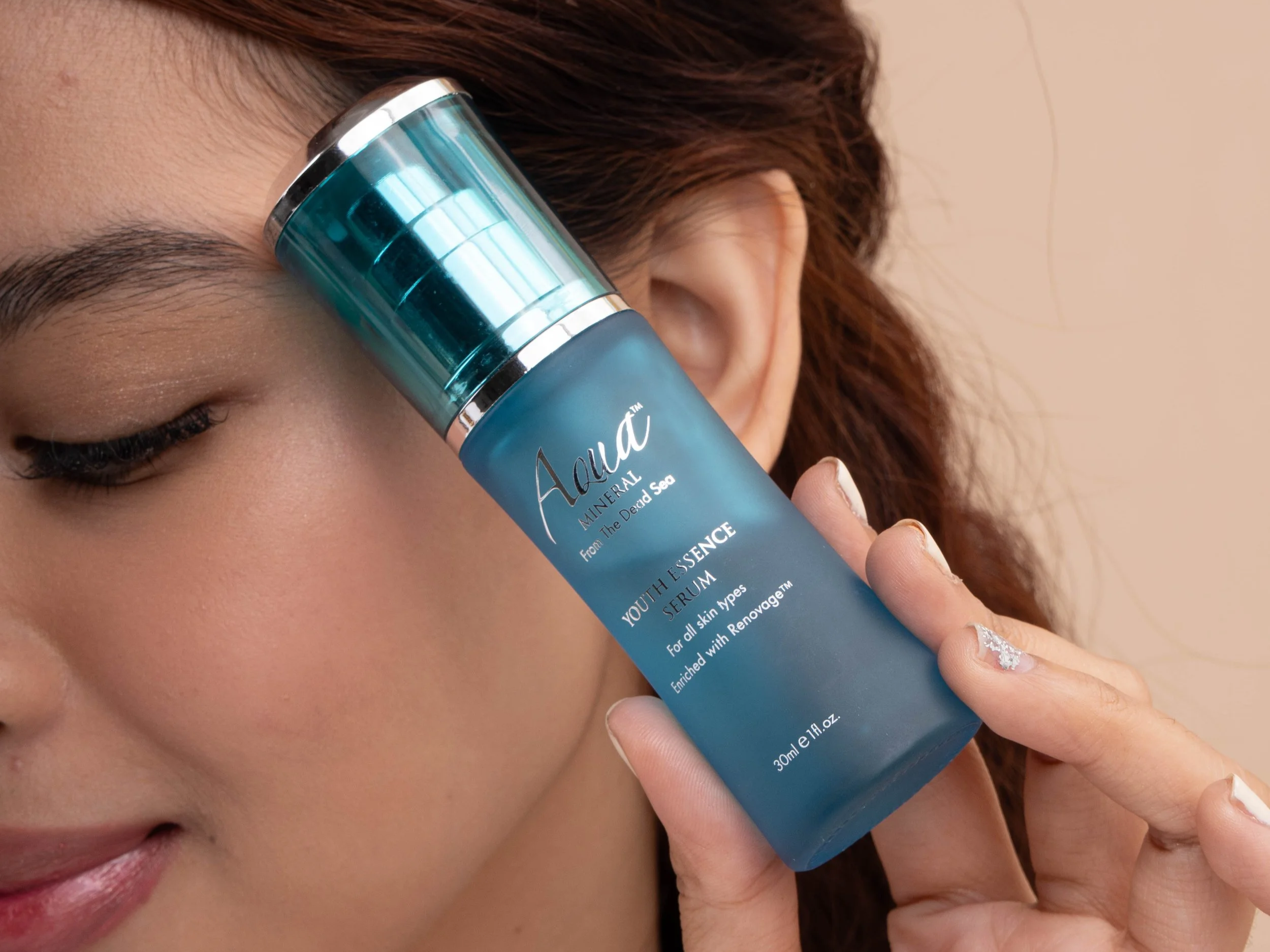 Close-up of a woman holding the blue Aqua Mineral Youth Essence Serum bottle against her cheek, with the product label and silver cap clearly visible.