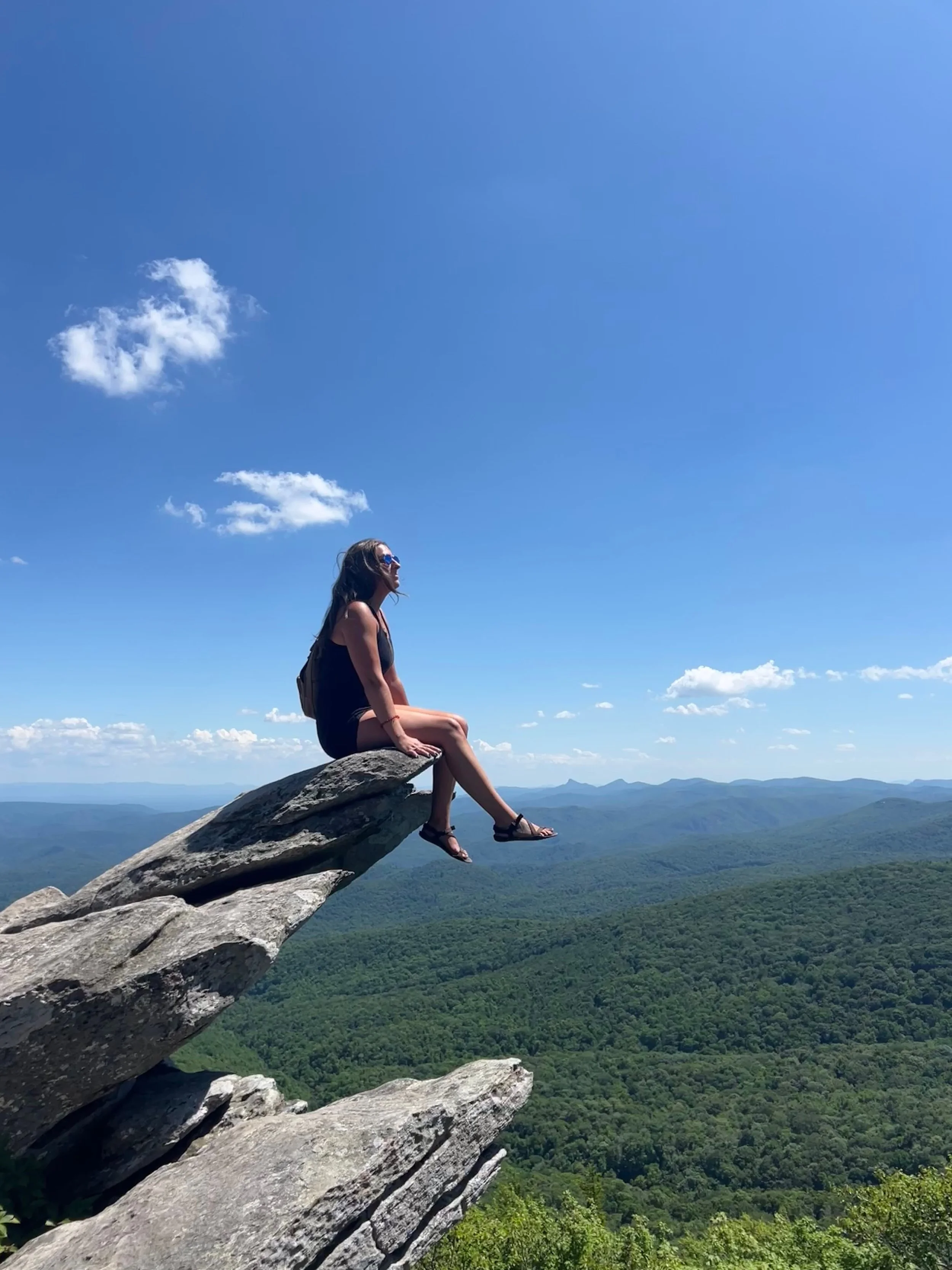 A woman sitting on a large rock formation on a mountain with a scenic view of green hills and blue sky with scattered clouds.